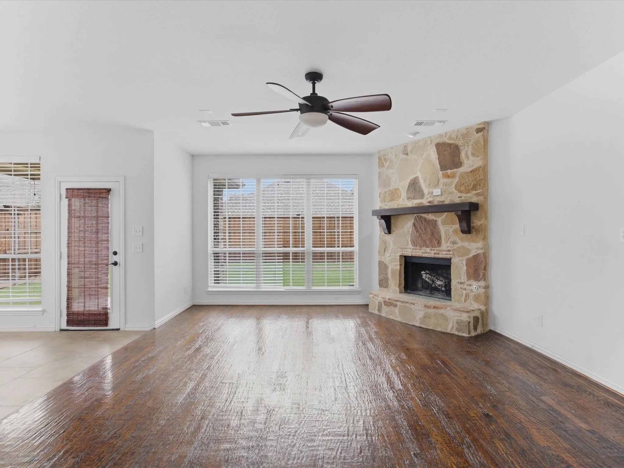 Spacious living area featuring wood-finish flooring, a stone fireplace with a wood mantel, multiple windows with blinds, a ceiling fan, and white walls