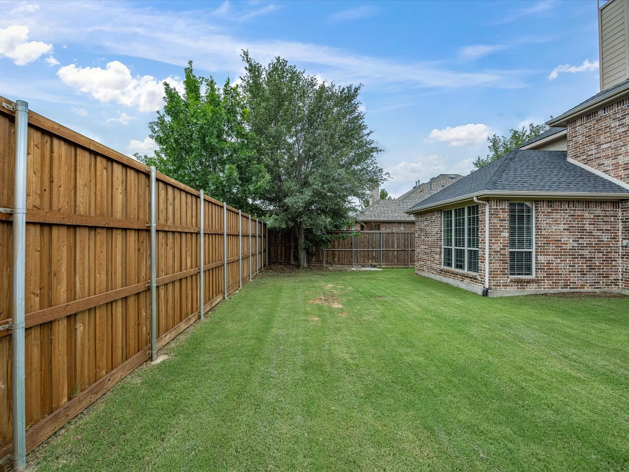 Expansive backyard featuring a wood privacy fence with metal posts, a brick exterior, a dark shingle roof, and mature trees