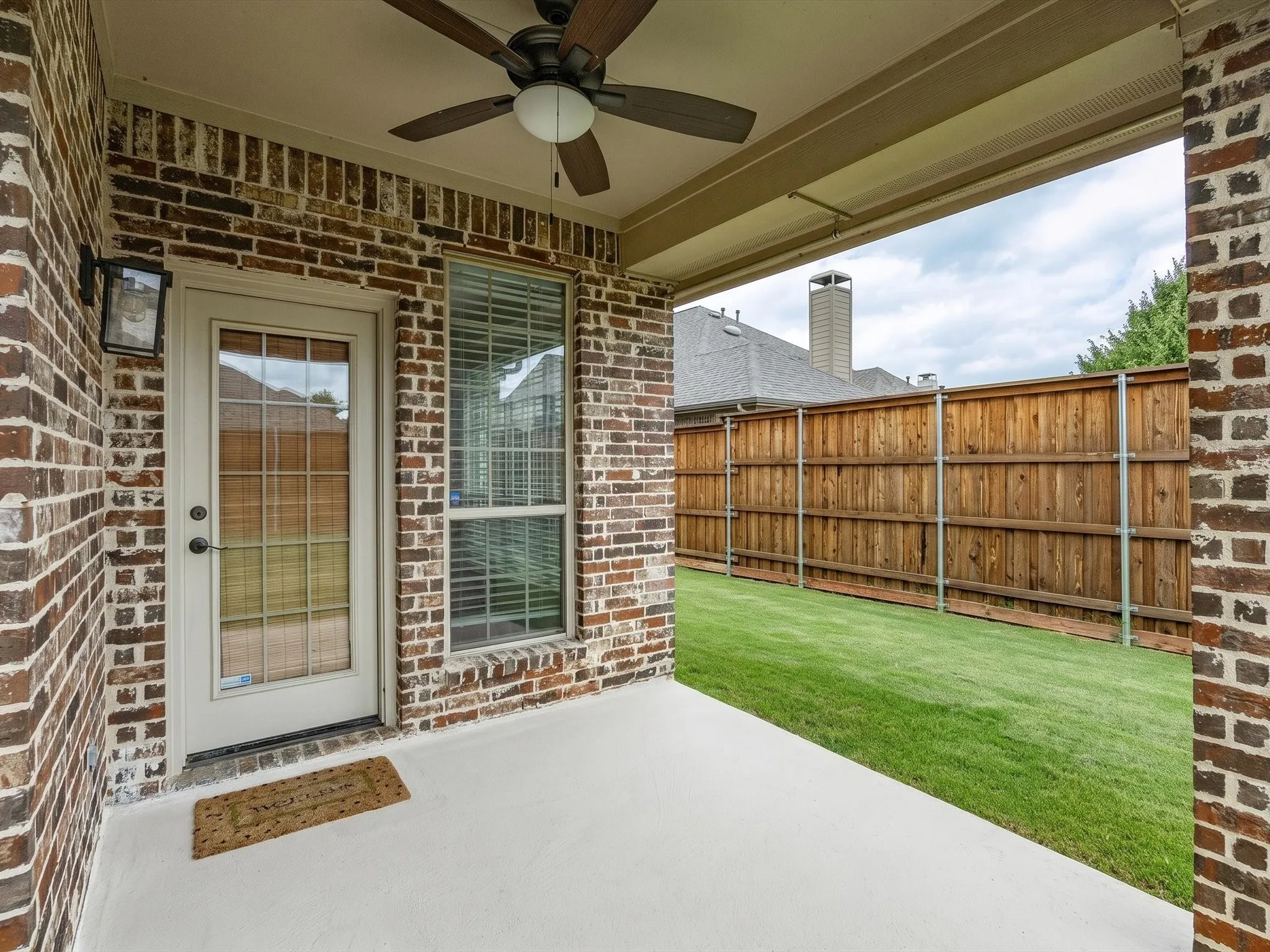 Covered outdoor patio with a ceiling fan, brick exterior, and a wood privacy fence bordering a grass yard