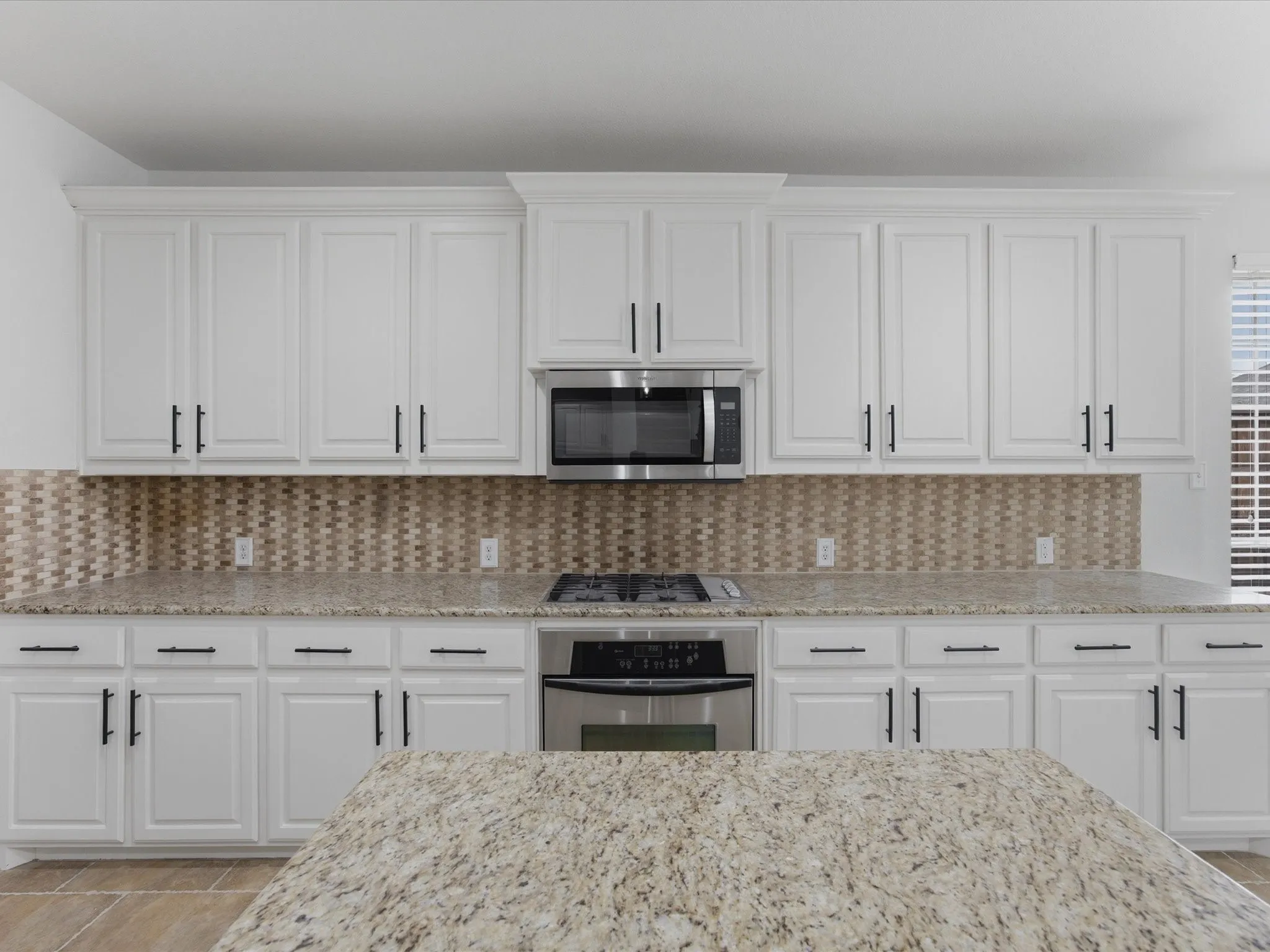 Kitchen featuring white shaker-style cabinetry with crown molding, light-toned granite countertops, a mosaic tile backsplash, stainless steel appliances, and a built-in gas cooktop