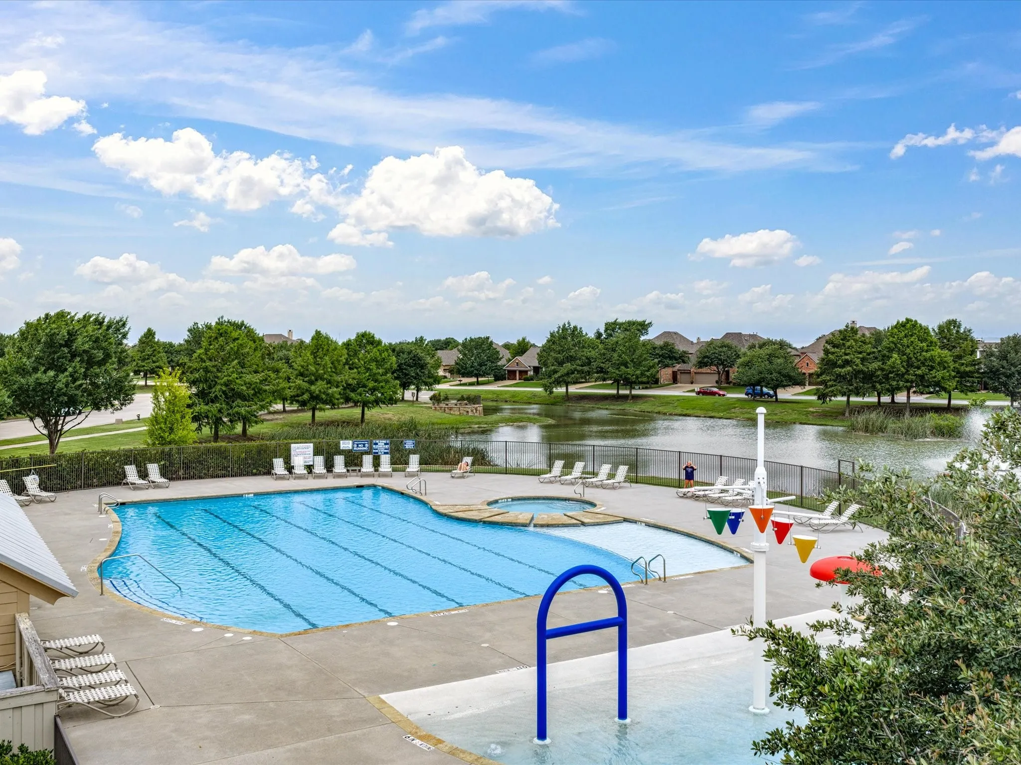 Community pool with lap lanes, an integrated spa, a splash pad area, and surrounding lounge seating, all situated alongside a scenic pond