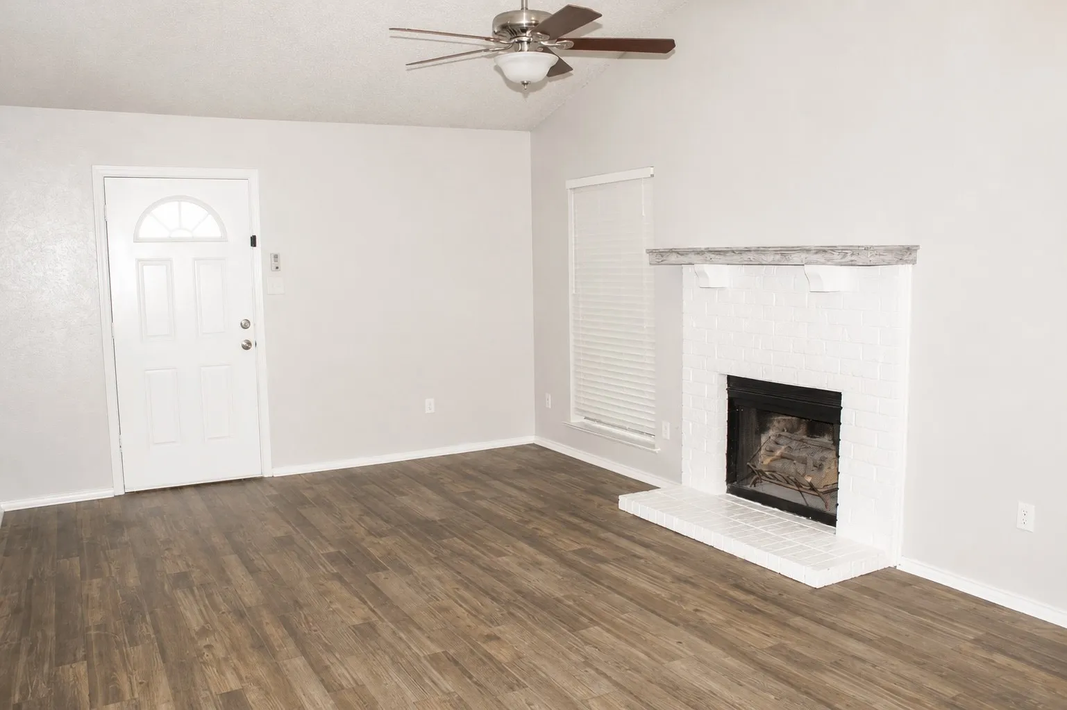 Spacious interior featuring wood-finish flooring, a white brick fireplace with mantel, white paneled entry door with arched transom, and a ceiling fan