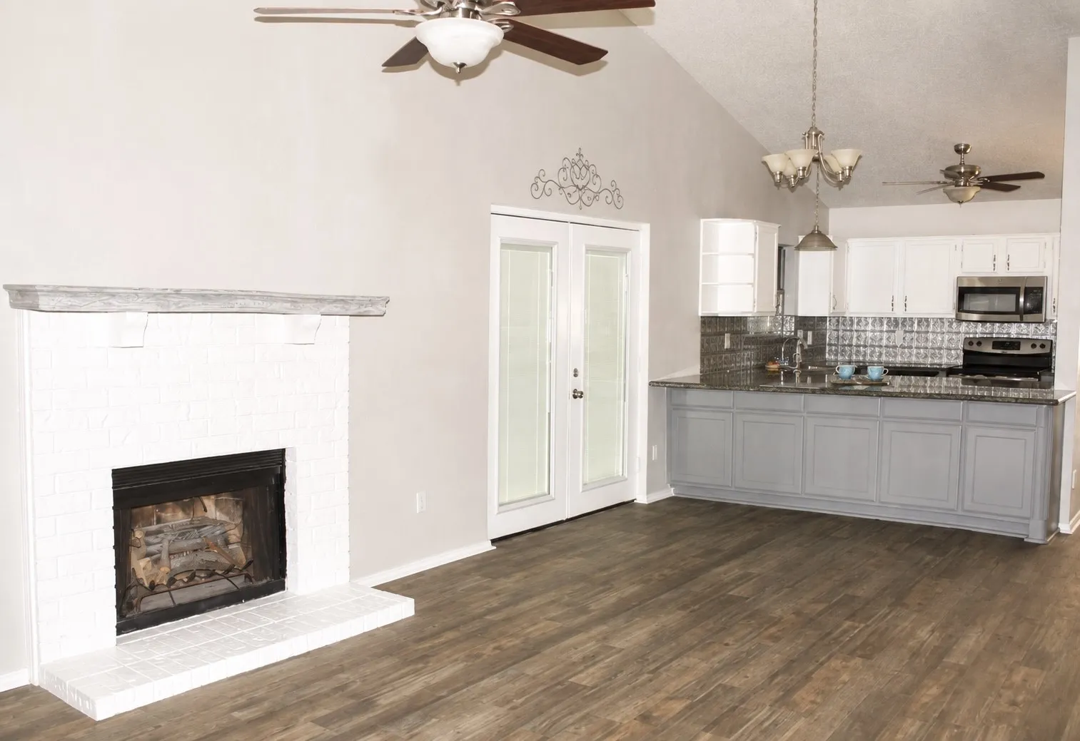 Wood-finish flooring throughout, vaulted ceiling, white brick fireplace, French doors, and a kitchen with white upper and grey lower cabinetry