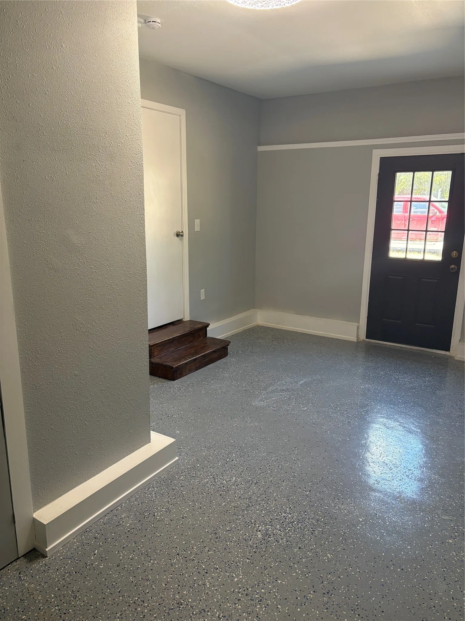 Textured gray walls with white trim, speckled epoxy flooring, a dark paneled door with glass inserts, and a white interior door with wood-finish steps