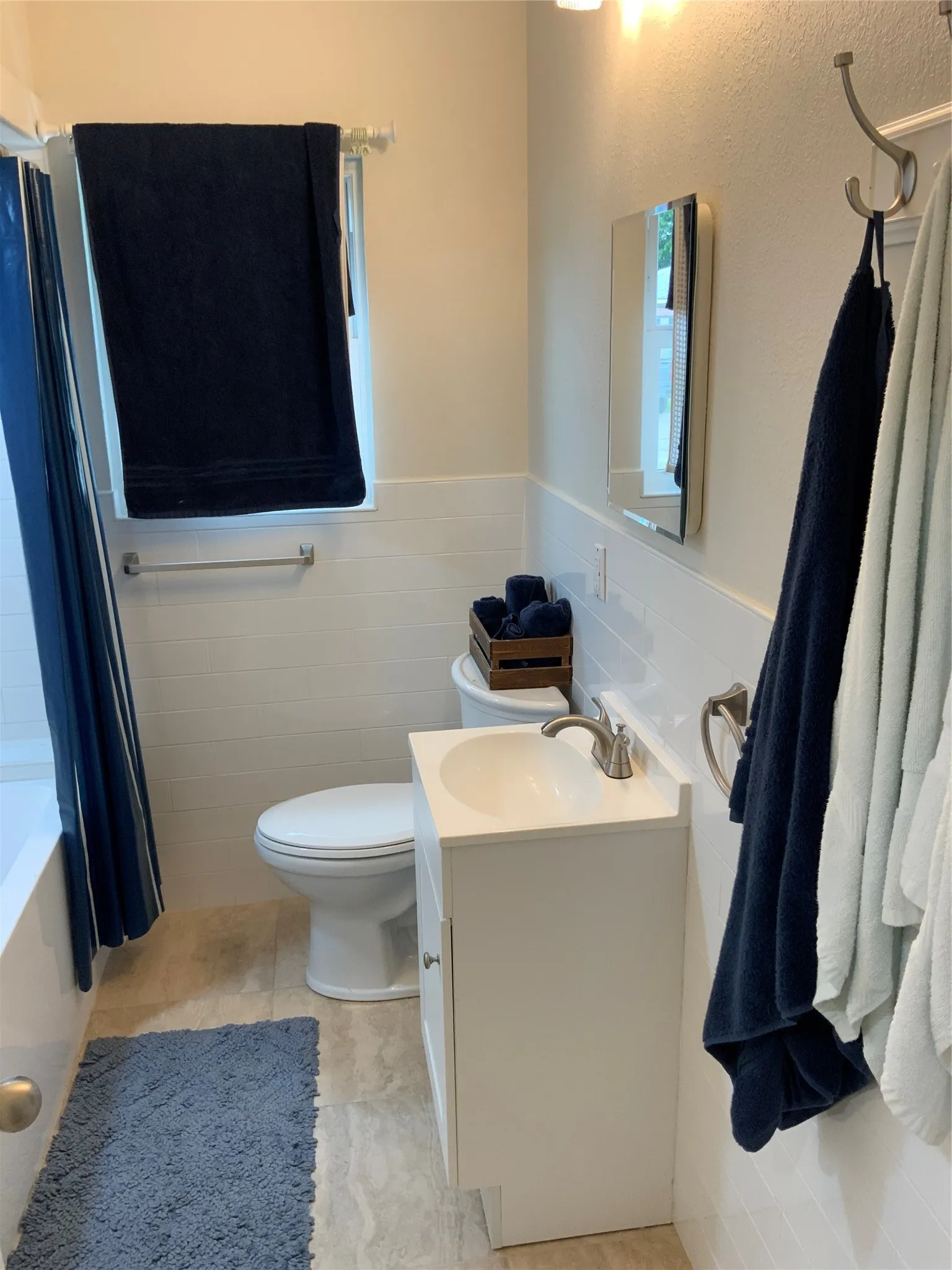 Compact bathroom featuring a white vanity with integrated sink, white subway tile wainscoting, a toilet, a standard bathtub with shower, and light-toned tile flooring