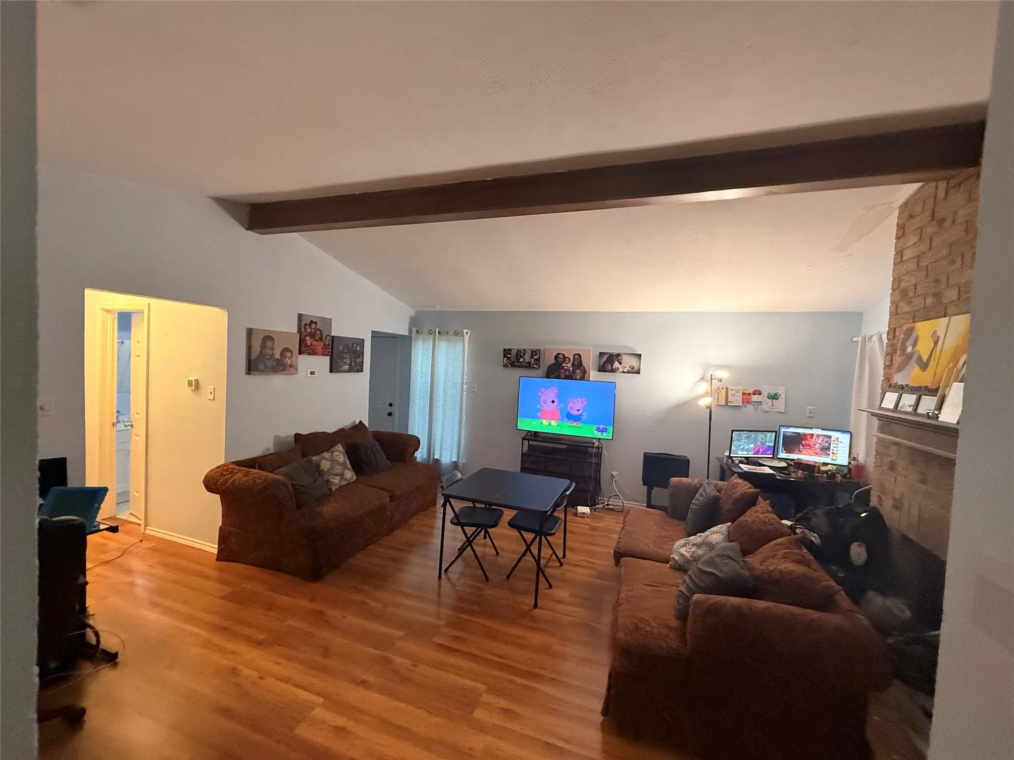 Spacious living area featuring wood-finish flooring, a vaulted ceiling with exposed wood beams, and a brick-faced fireplace with a mantel