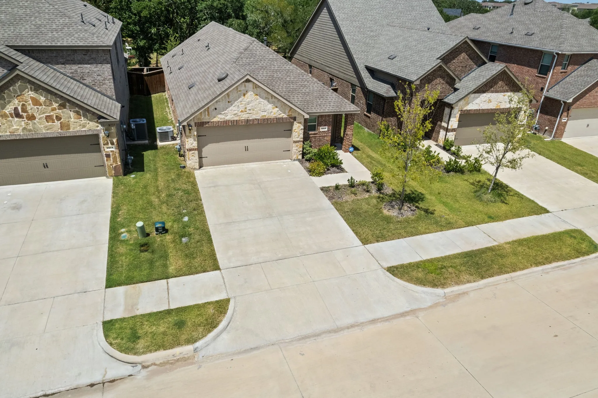 Aerial view of front of home featuring a stone and brick facade, a single-door two-car garage, a concrete driveway, and mature landscaping with established trees.
