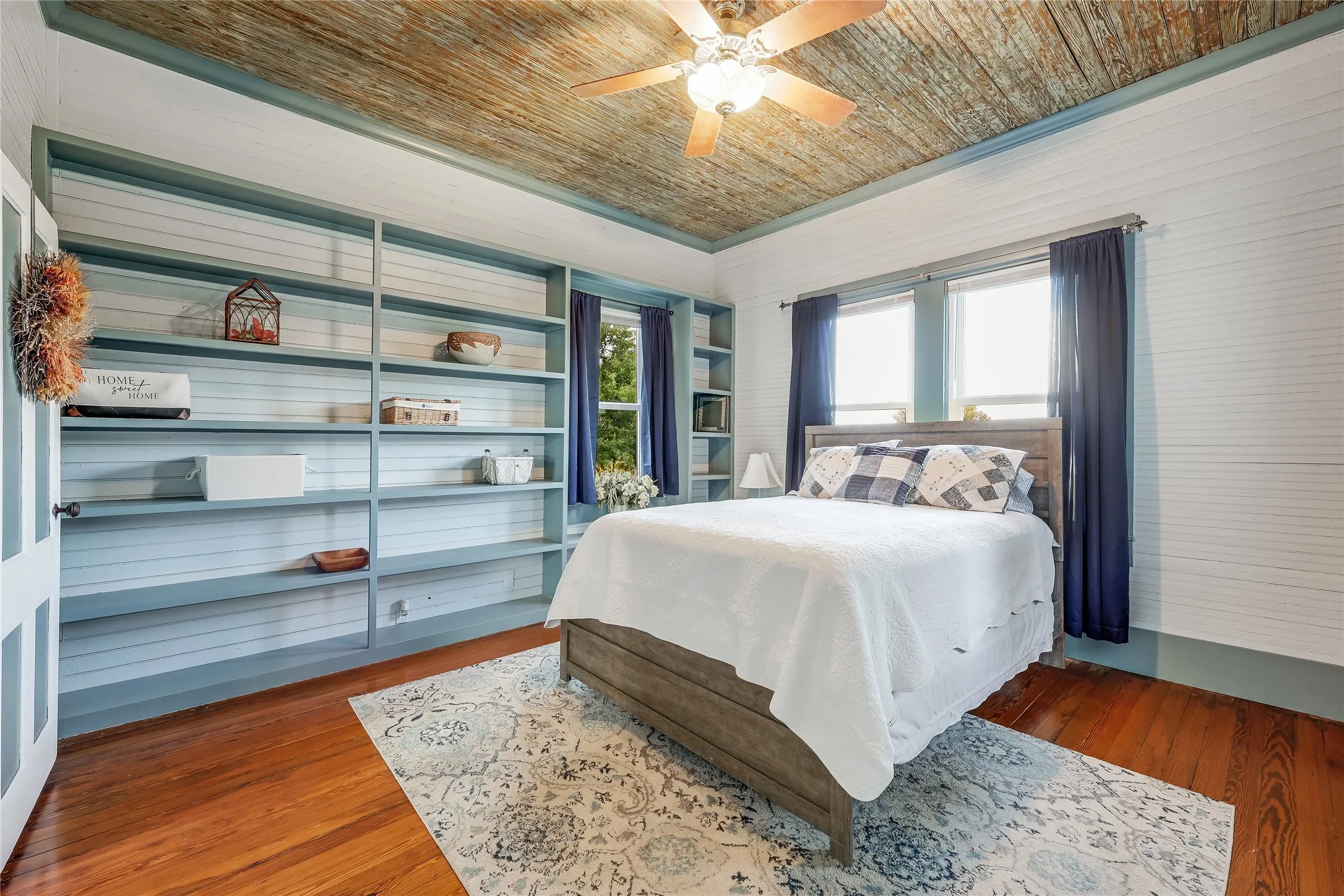 Room featuring a distressed wood-finish ceiling with a ceiling fan, built-in shelving, wood-finish flooring, and two windows