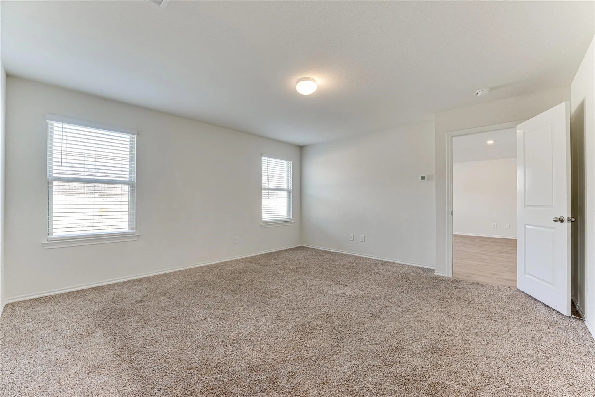 Spacious room featuring neutral carpeting, white walls, two windows with blinds, a ceiling-mounted light fixture, and an open doorway revealing wood-finish flooring in the adjacent room