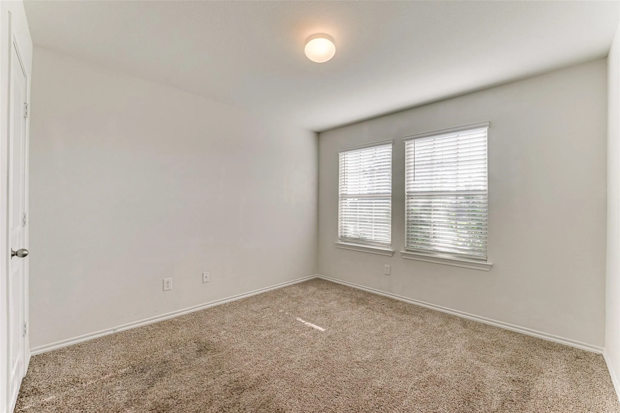 Carpeted interior room featuring two windows with blinds, a ceiling-mounted light fixture, white walls, and white baseboards