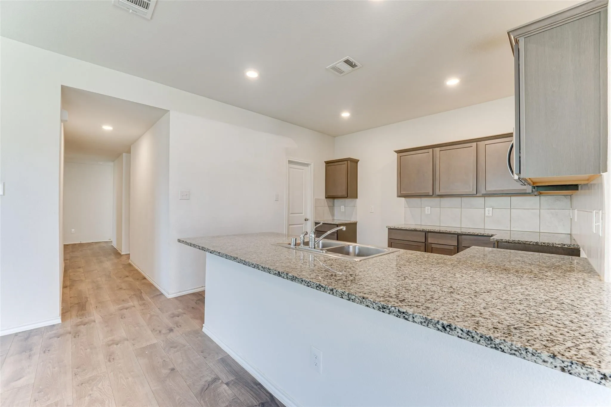 Kitchen featuring an expansive granite countertop island with an undermount double-basin sink, light wood-finish flooring, recessed lighting, and grey cabinetry with a tile backsplash