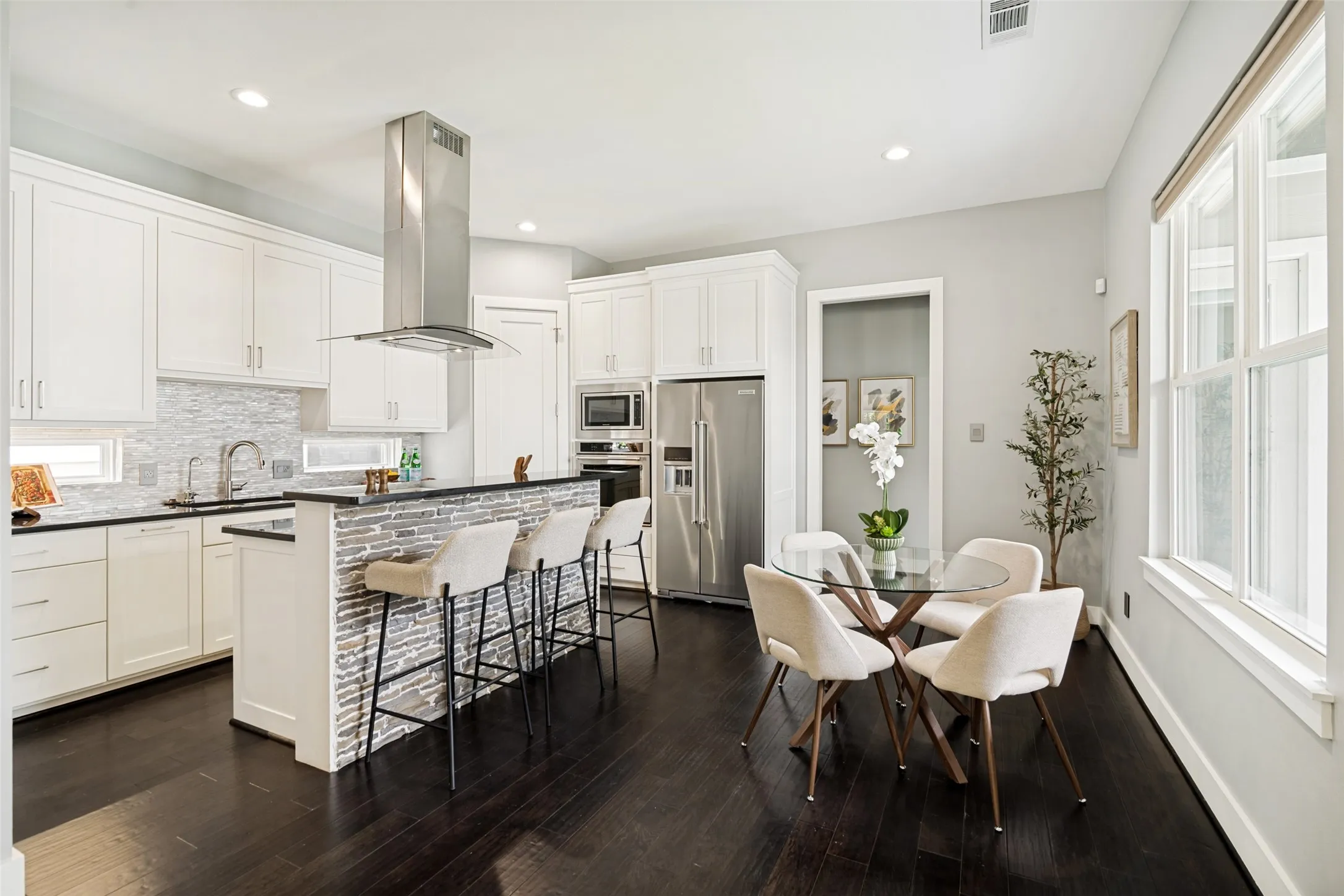 Light-filled kitchen and breakfast room with rectangular windows, island with breakfast bar, commercial-style vent hood, white stone backsplash, and spacious walk-in pantry.