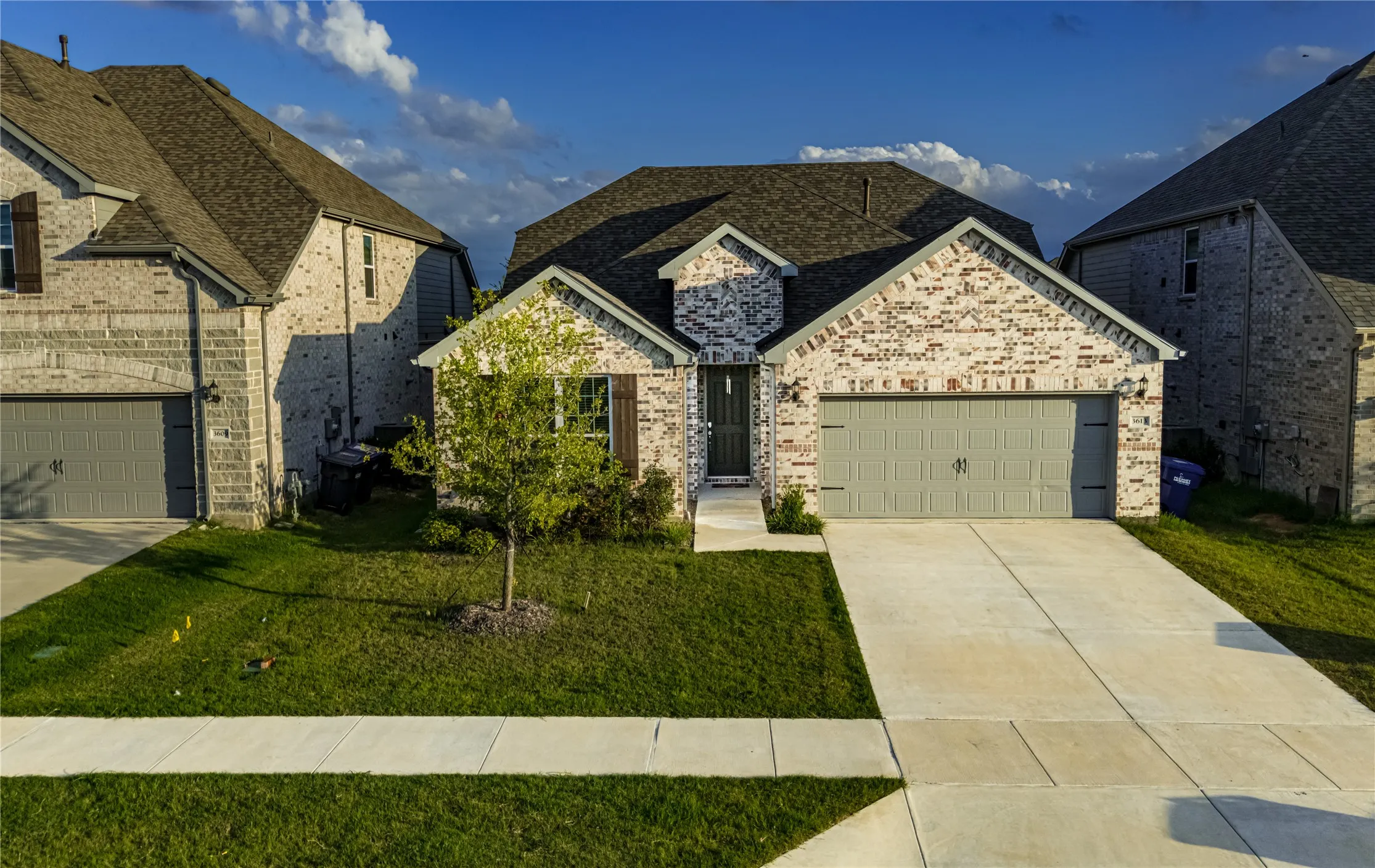 Brick facade home featuring an attached two-car garage, concrete driveway, and front yard with a young tree