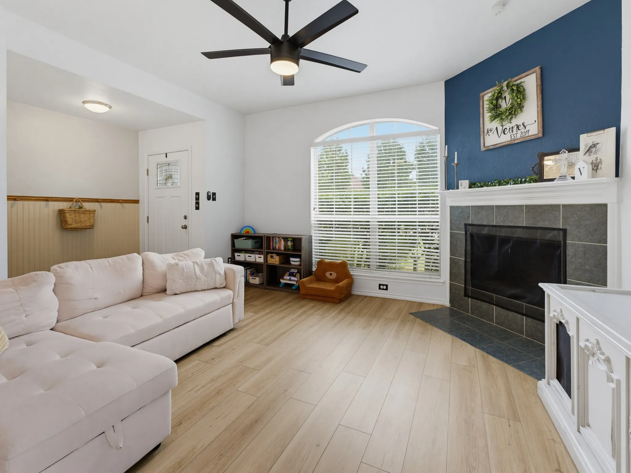 Living area featuring wood-finish flooring, a contemporary fireplace with a tiled surround and hearth, and a large arched window with horizontal blinds