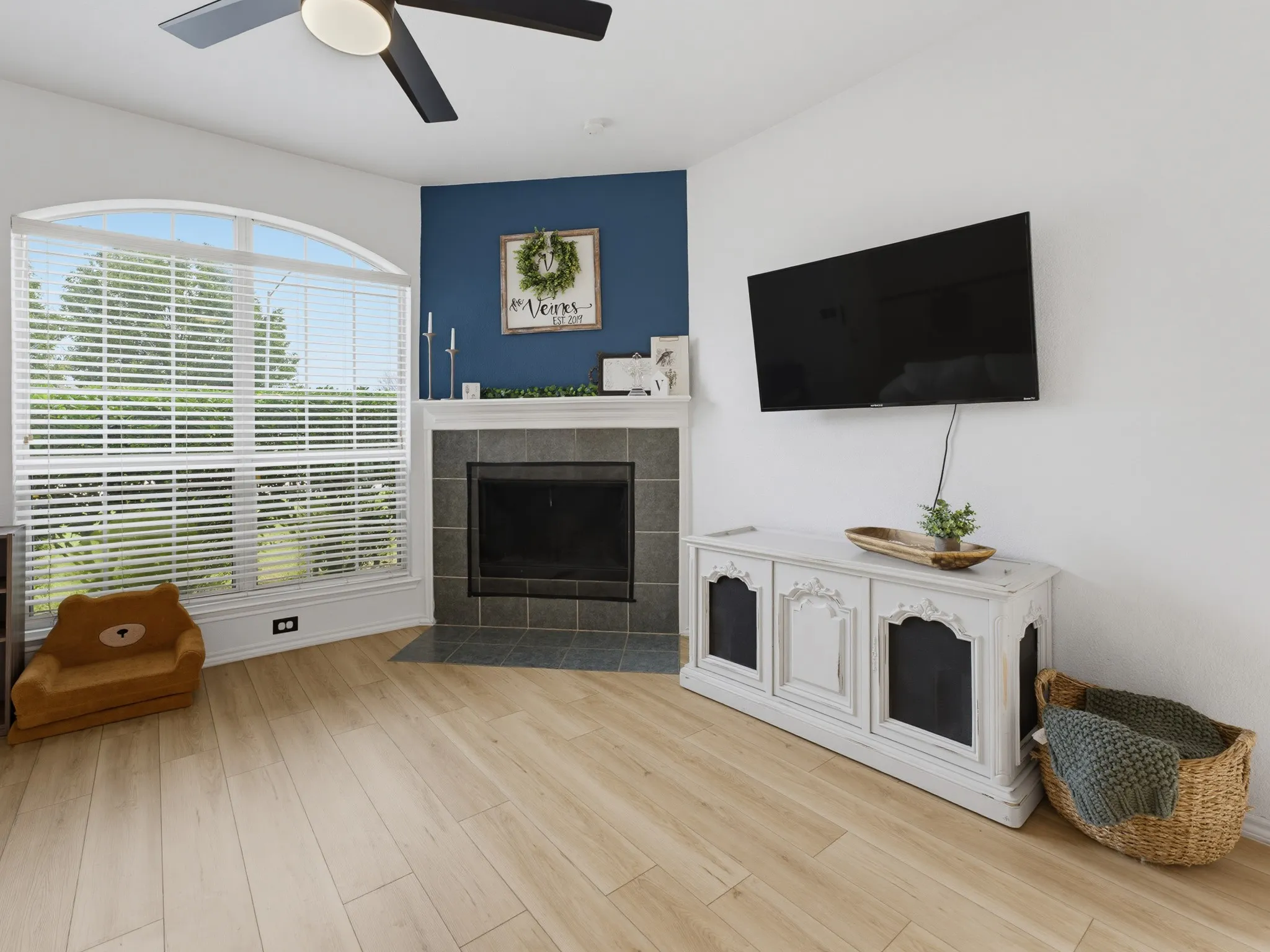 Living area with wood-finish flooring, a ceiling fan with integrated lighting, and a corner fireplace featuring a white mantle and dark tile surround