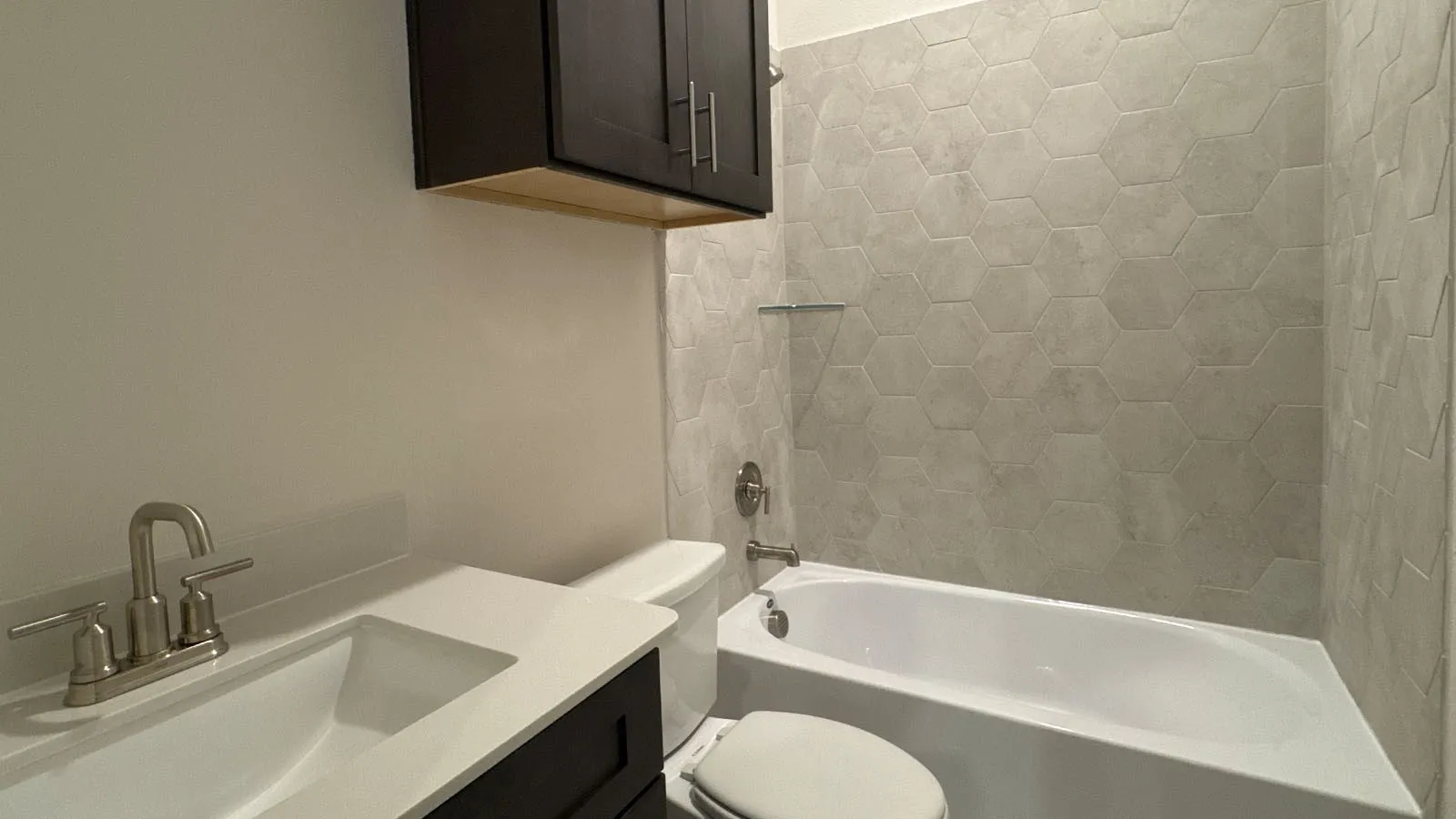 Built-in white bathtub with hexagonal tile surround, dark wood-finish cabinetry, white countertop, and brushed nickel fixtures