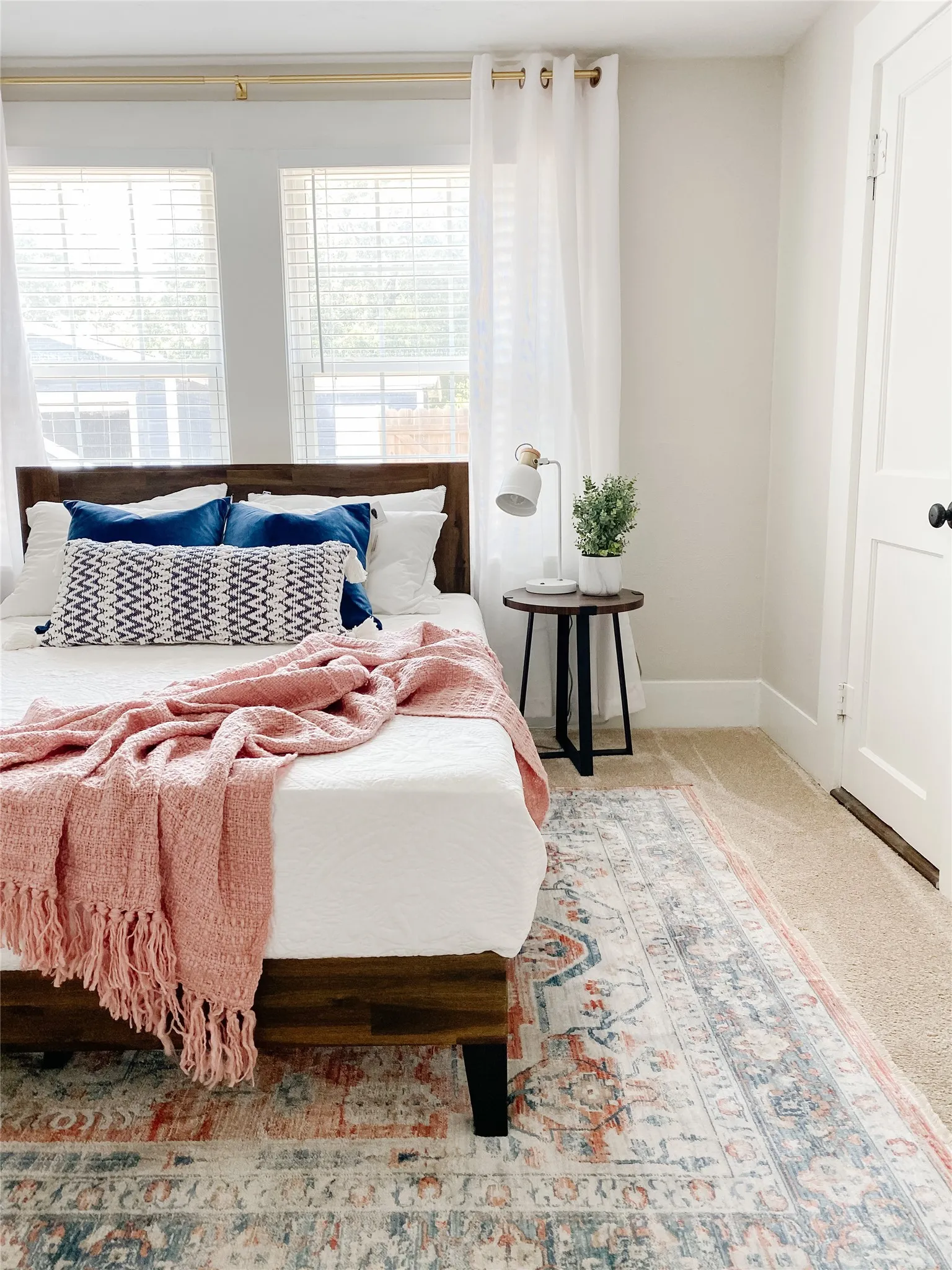 Warm wood-tone bed frame in a room featuring neutral wall tones, two paneled windows with blinds, and a white interior door with black hardware