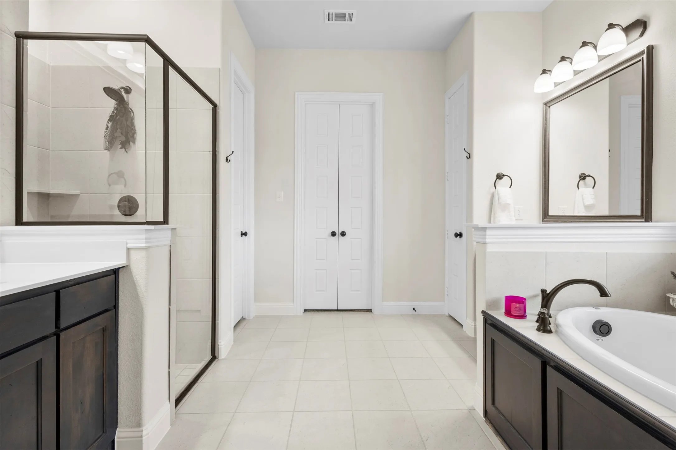 Spacious bathroom featuring light-toned tile flooring, a dark wood-finish vanity with a white countertop, a walk-in shower with a built-in bench and dark-framed glass enclosure, a white soaking tub with dark bronze fixtures, and a mirrored vanity with a five-light fixture