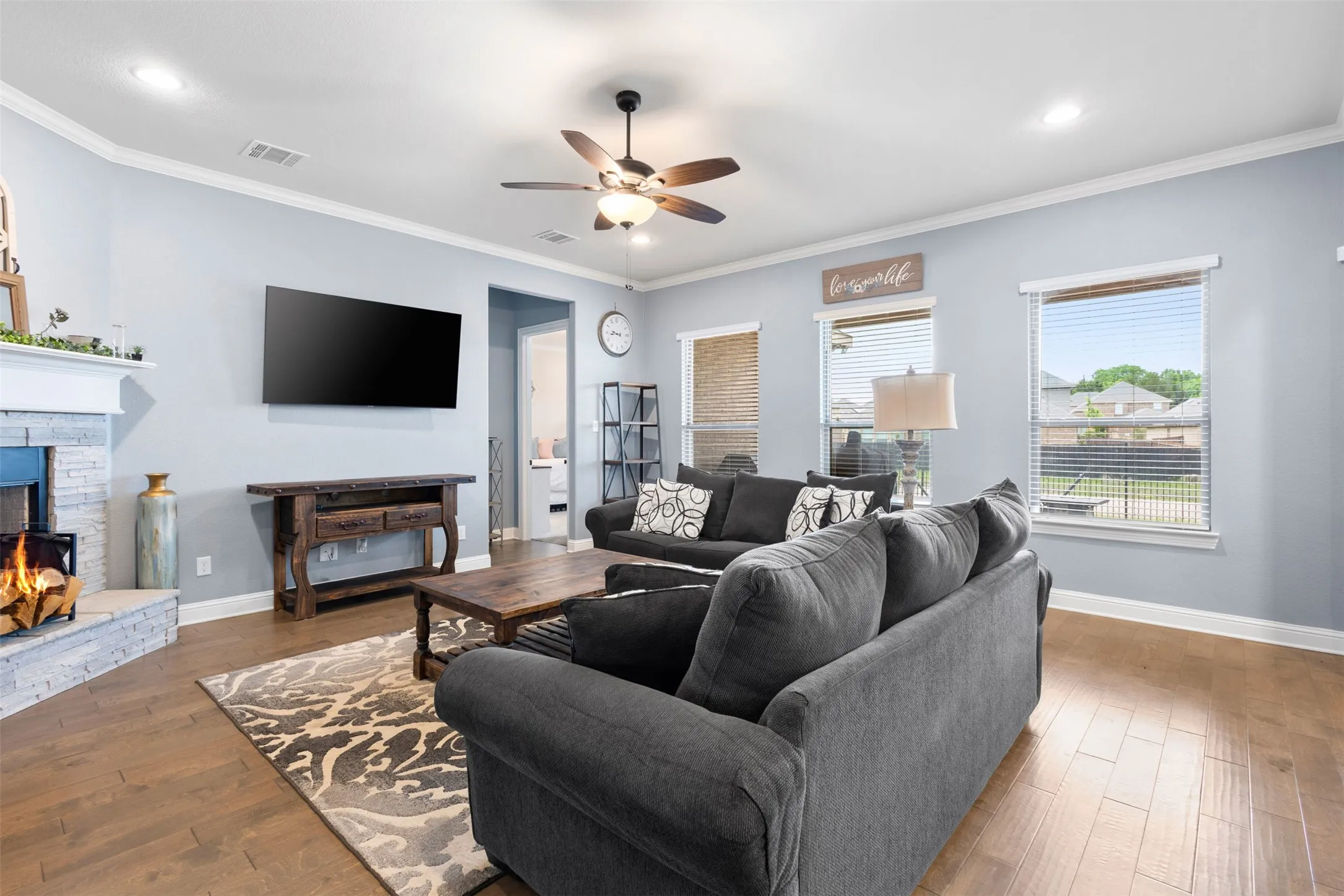 Living area featuring wood-finish flooring, a stone-clad fireplace, crown molding, a ceiling fan, and recessed lighting