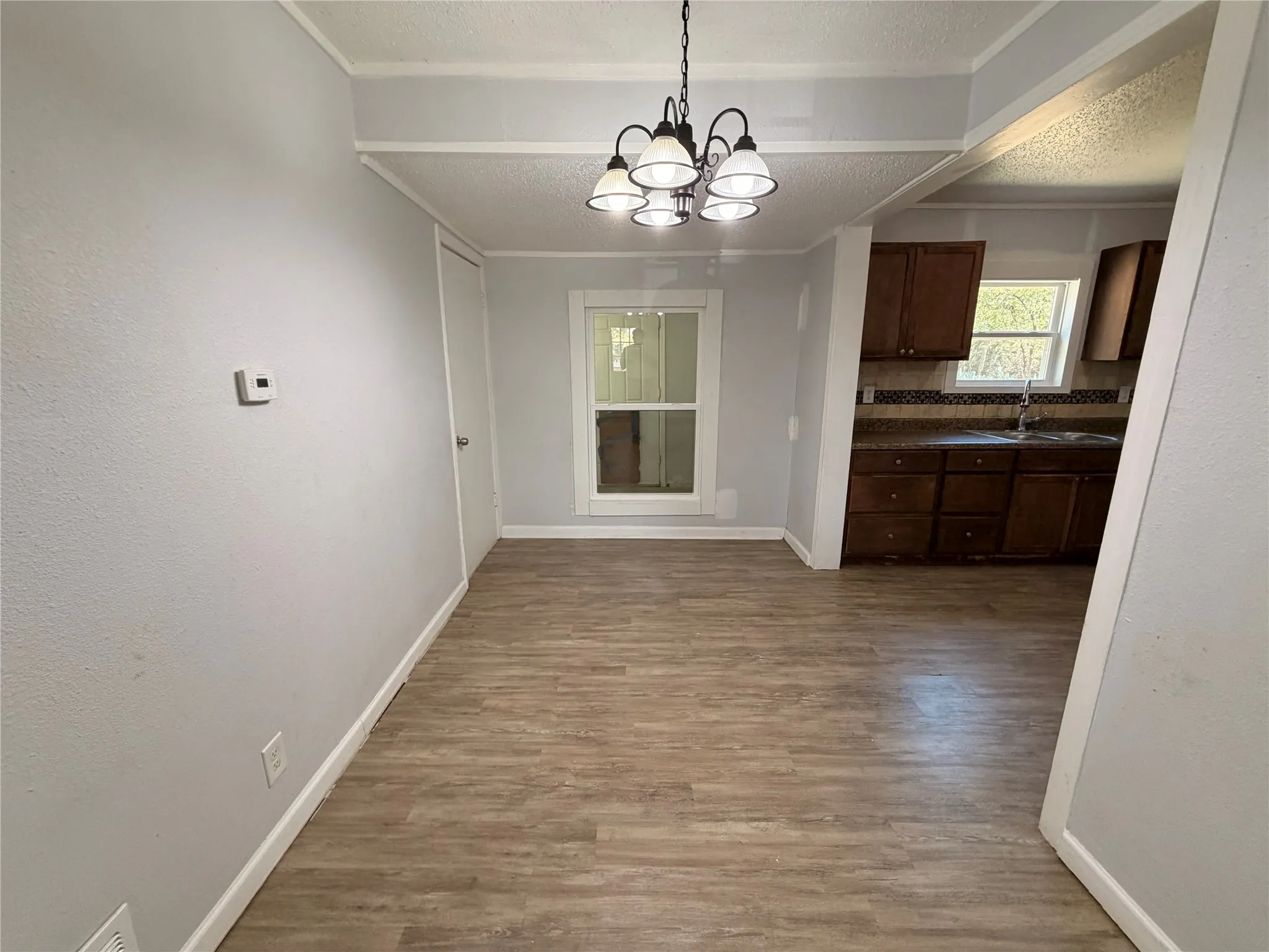 Wood-finish flooring throughout, with a classic chandelier and an open doorway to the kitchen featuring wood cabinetry and a tiled backsplash