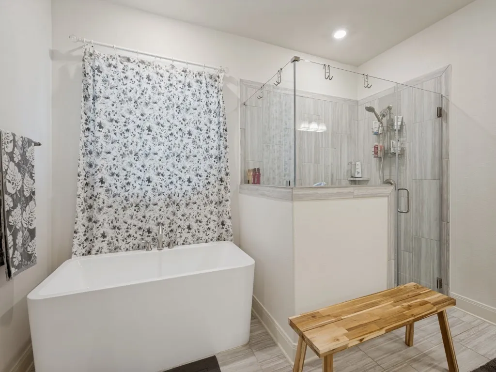 Spa-inspired primary bath featuring a freestanding soaking tub, oversized glass-enclosed shower, elegant tile finishes, and a bright, relaxing atmosphere. The curtain behind the tub is for privacy. Frameless glass shower has a sprayhead as well as a handheld sprayer that is such a help after cleaning the shower! Wood-look tile floors, recessed lighting, tall baseboards all add to the elegance of the primary bath.