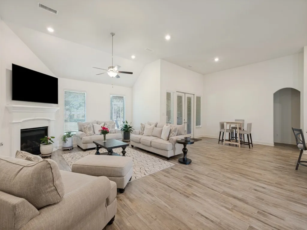 The bright, open living room with soaring ceilings, wood-look tile flooring, and abundant natural light is an inviting space for everyday living and entertaining. Beautiful wood-look tile floors, focal point fireplace, lots of recessed lighting.  The arched doorway to the right leads to the primary suite