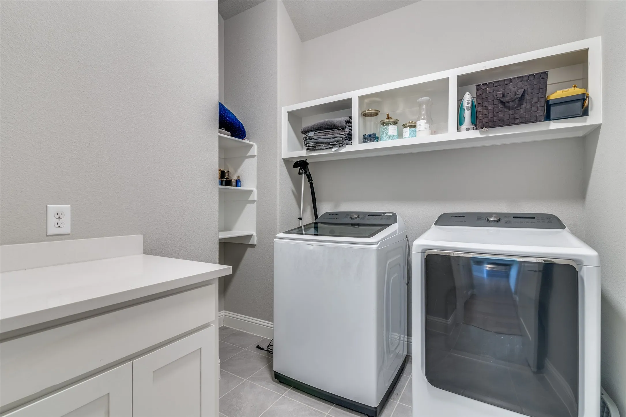 Dedicated laundry area featuring a white countertop with integrated cabinetry, built-in shelving, overhead open storage, and tile flooring