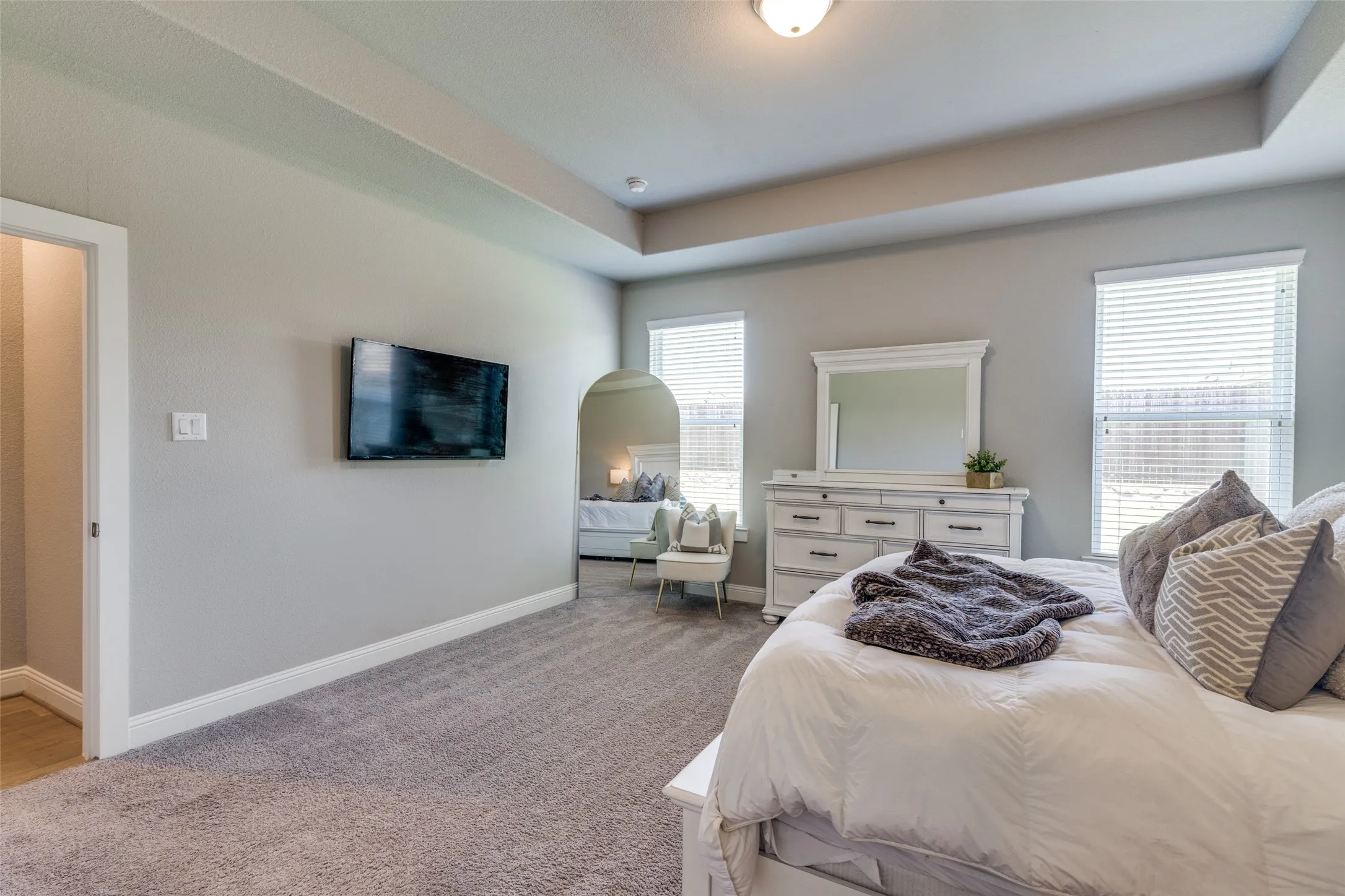 Spacious bedroom featuring light gray walls, plush gray carpet, a tray ceiling, two windows with blinds, and a doorway leading to an additional space