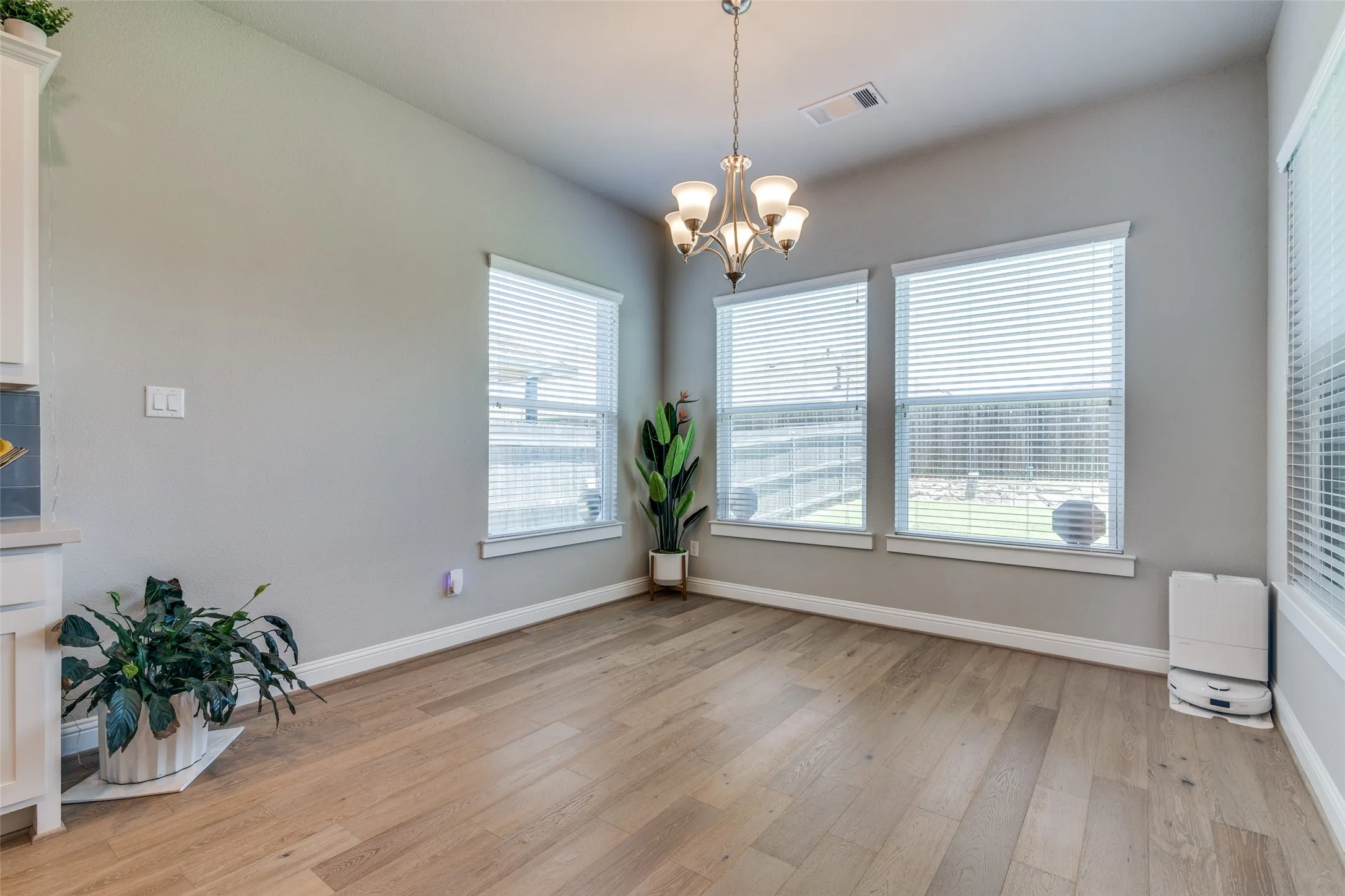 Spacious dining area featuring wood-finish flooring, a contemporary chandelier, and three large windows with horizontal blinds