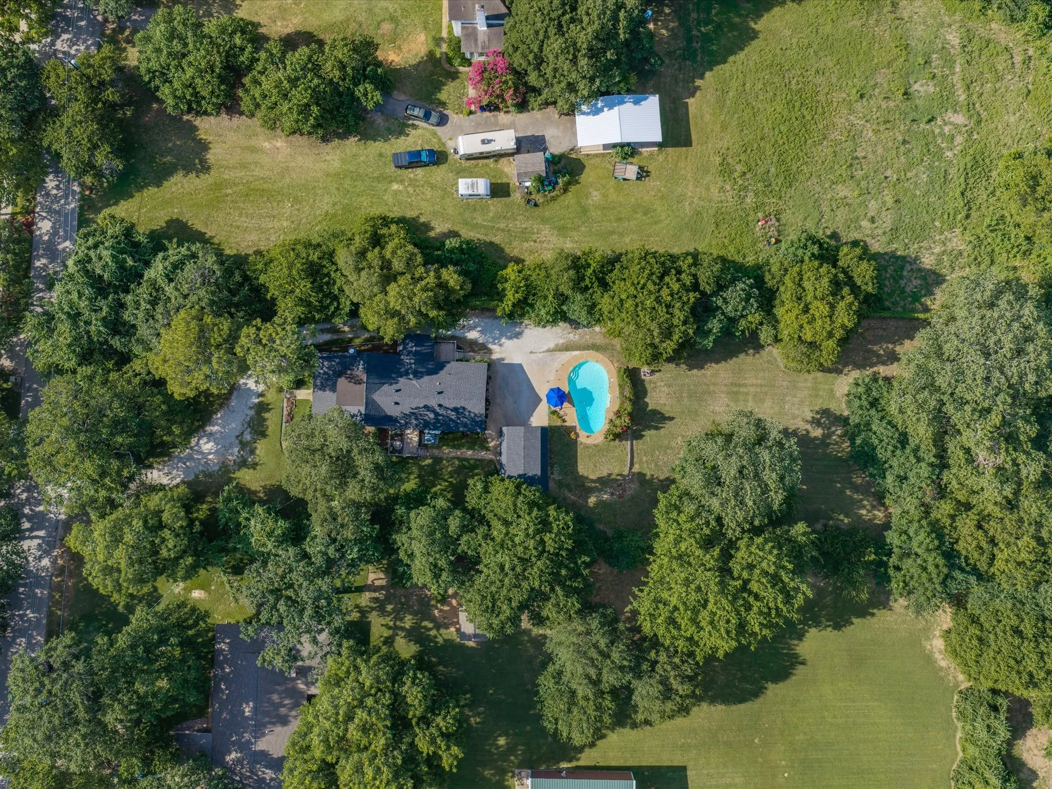 Aerial perspective showcasing a property surrounded by mature trees, featuring a dark-toned roofline, a freeform swimming pool, and a gravel driveway