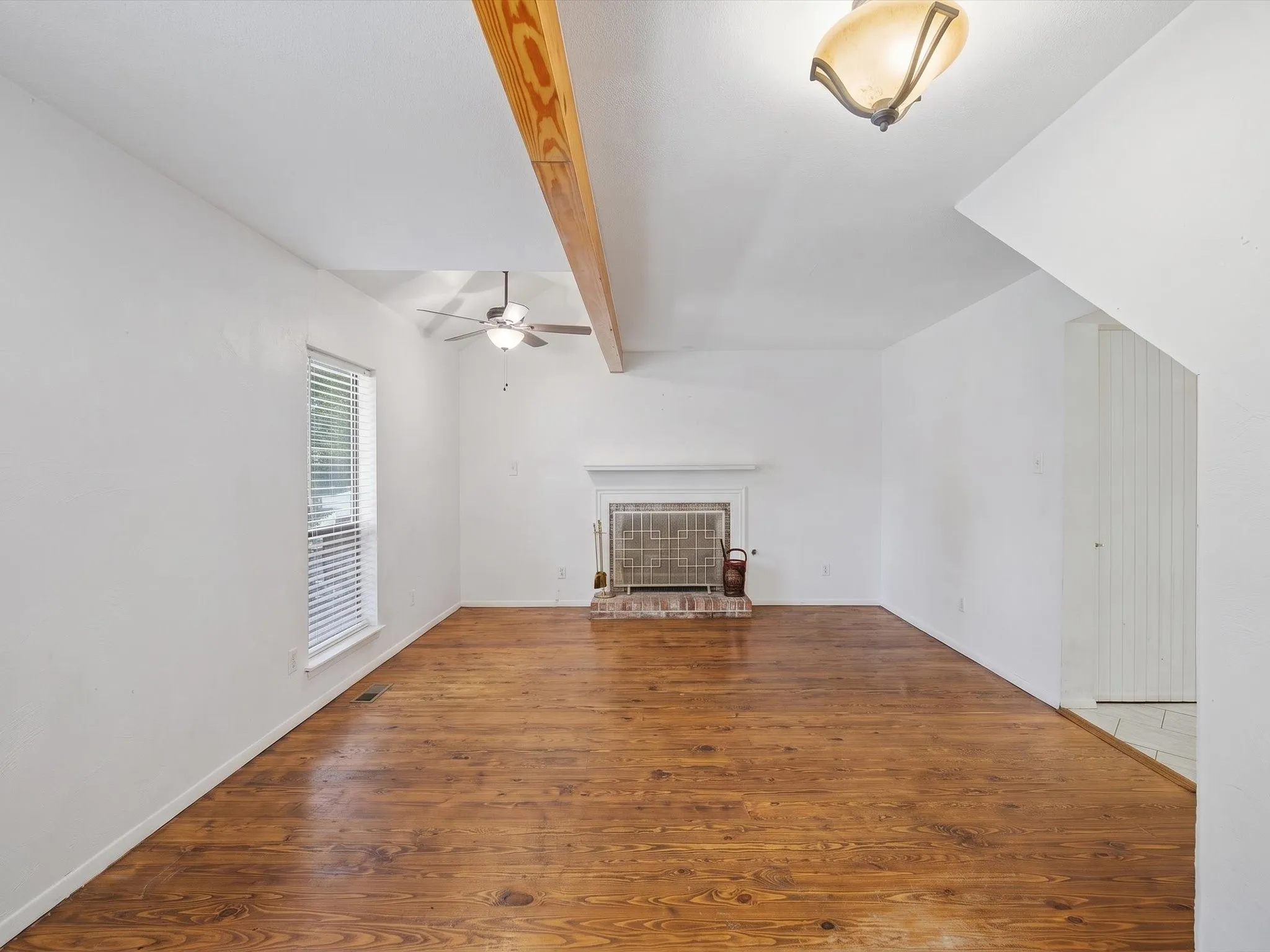 Spacious living area featuring wood-finish flooring, a traditional fireplace with a brick hearth, a ceiling fan, and a prominent exposed wood beam