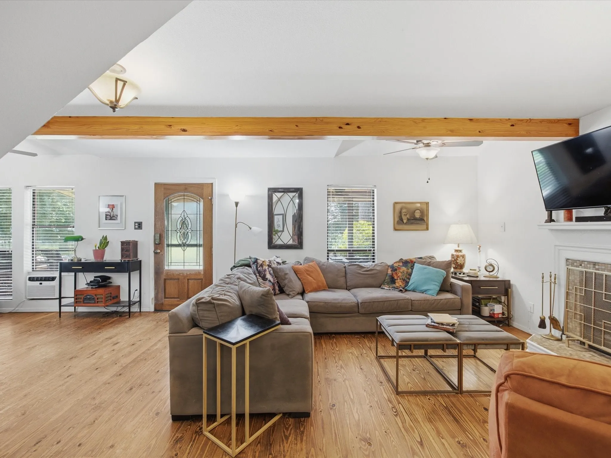 Spacious living area featuring wood-finish flooring, exposed ceiling beam, ceiling fan, and a decorative front entry door with leaded glass