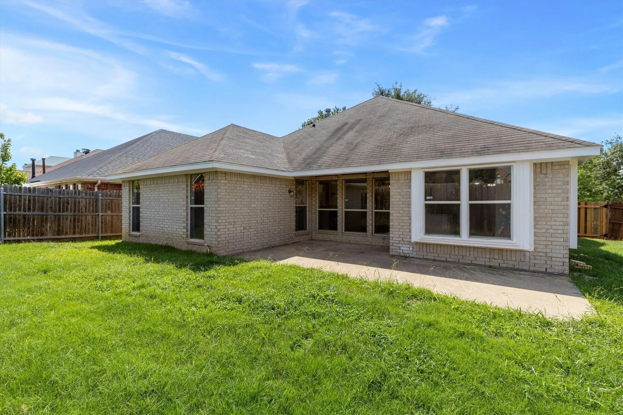 Expansive rear yard featuring a concrete patio, light-colored brick exterior, multiple windows, and a wood fence