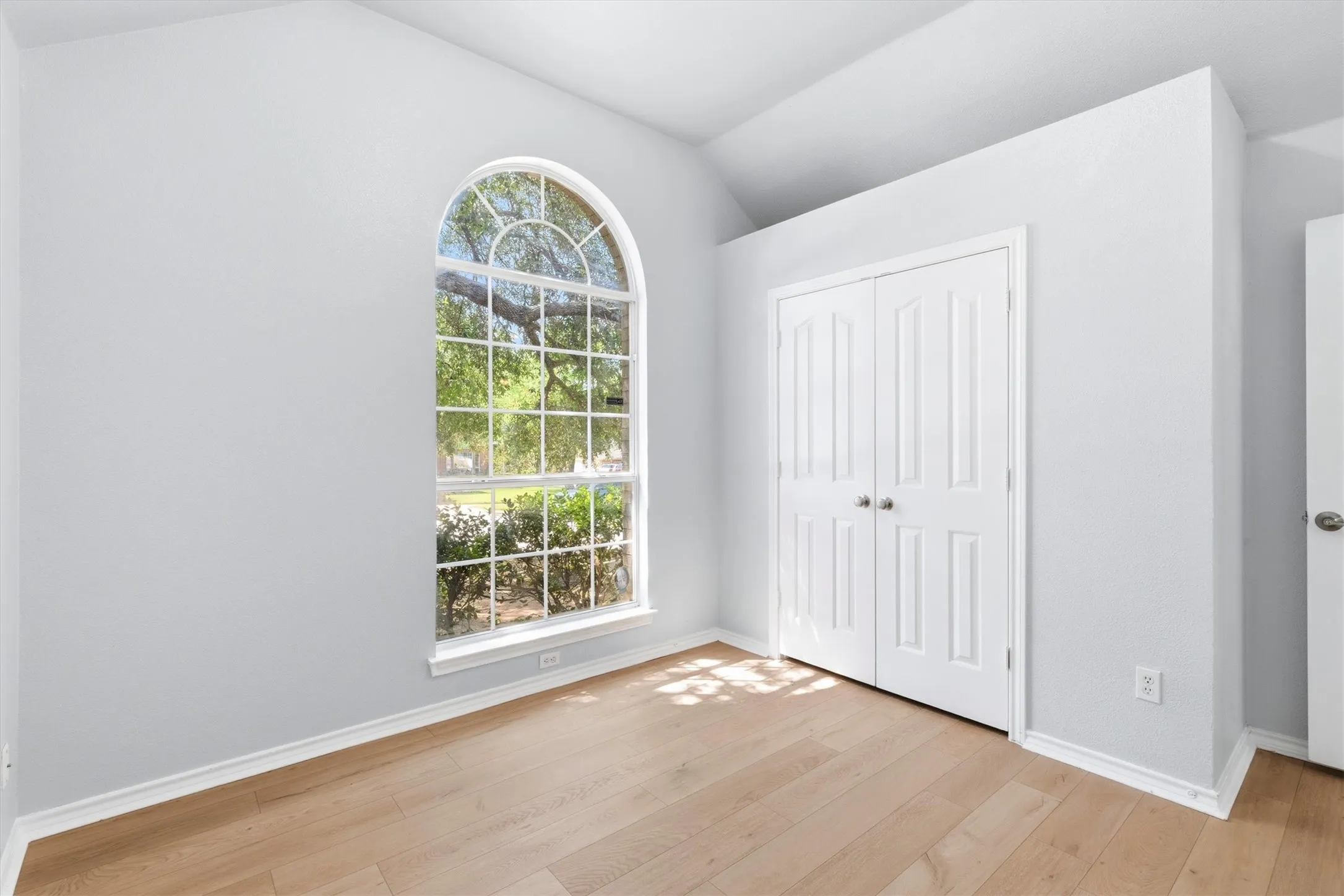 Light-filled interior featuring a large arched window, vaulted ceiling, wood-finish flooring, and paneled bifold closet doors