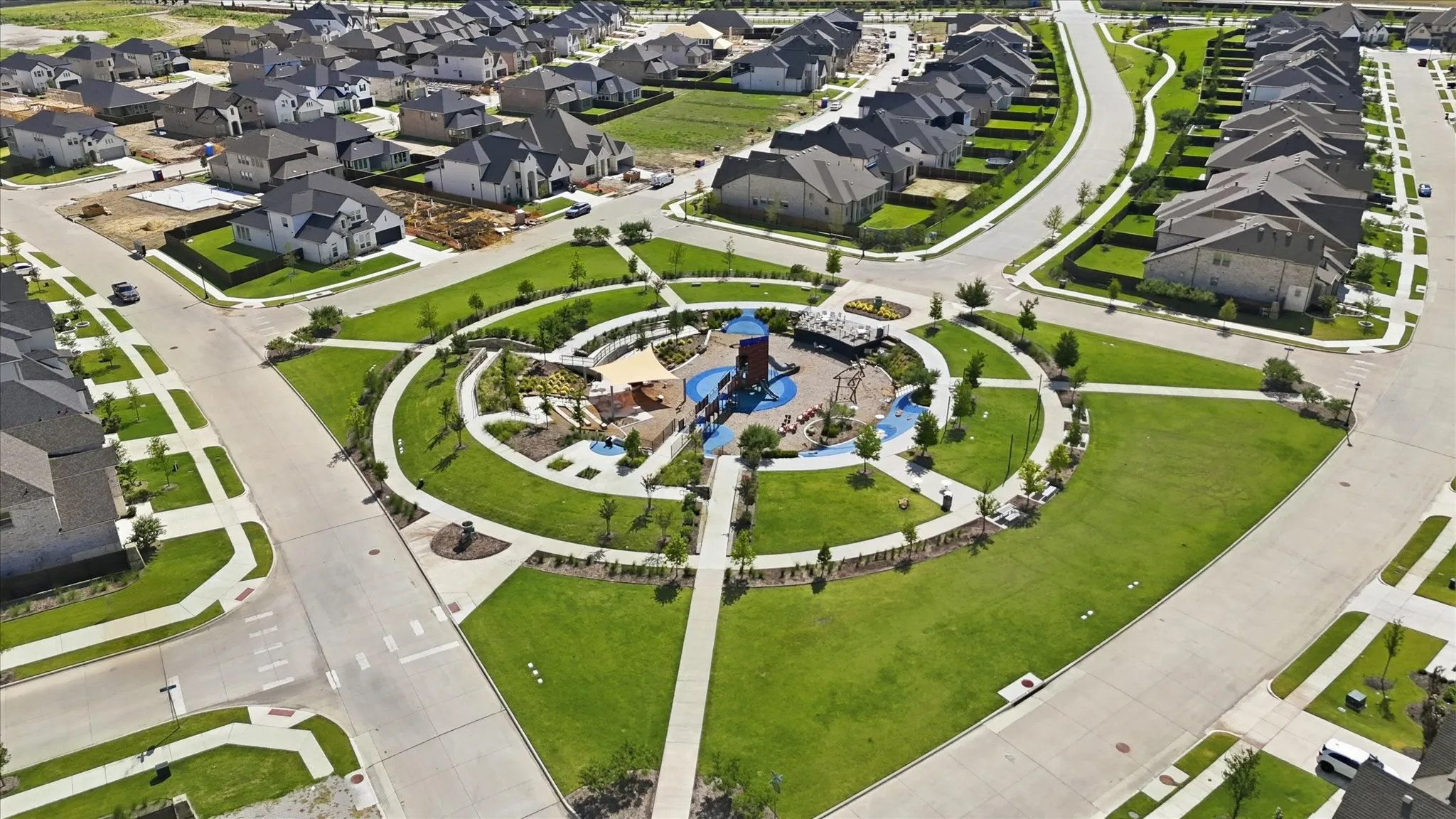 Central park area featuring a playground with shade structures, paved walking paths, and expansive green spaces