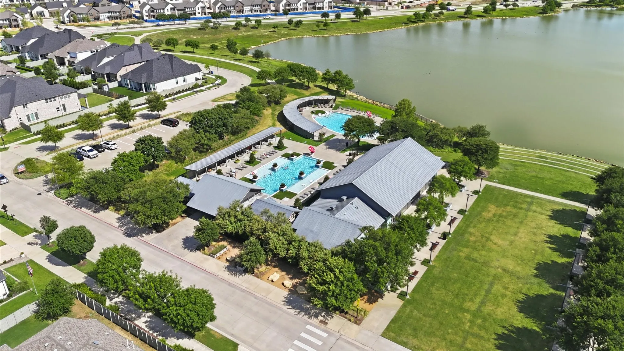 Community amenity center featuring two swimming pools, a clubhouse with a metal roof, an expansive lawn, and mature landscaping, all situated alongside a large body of water