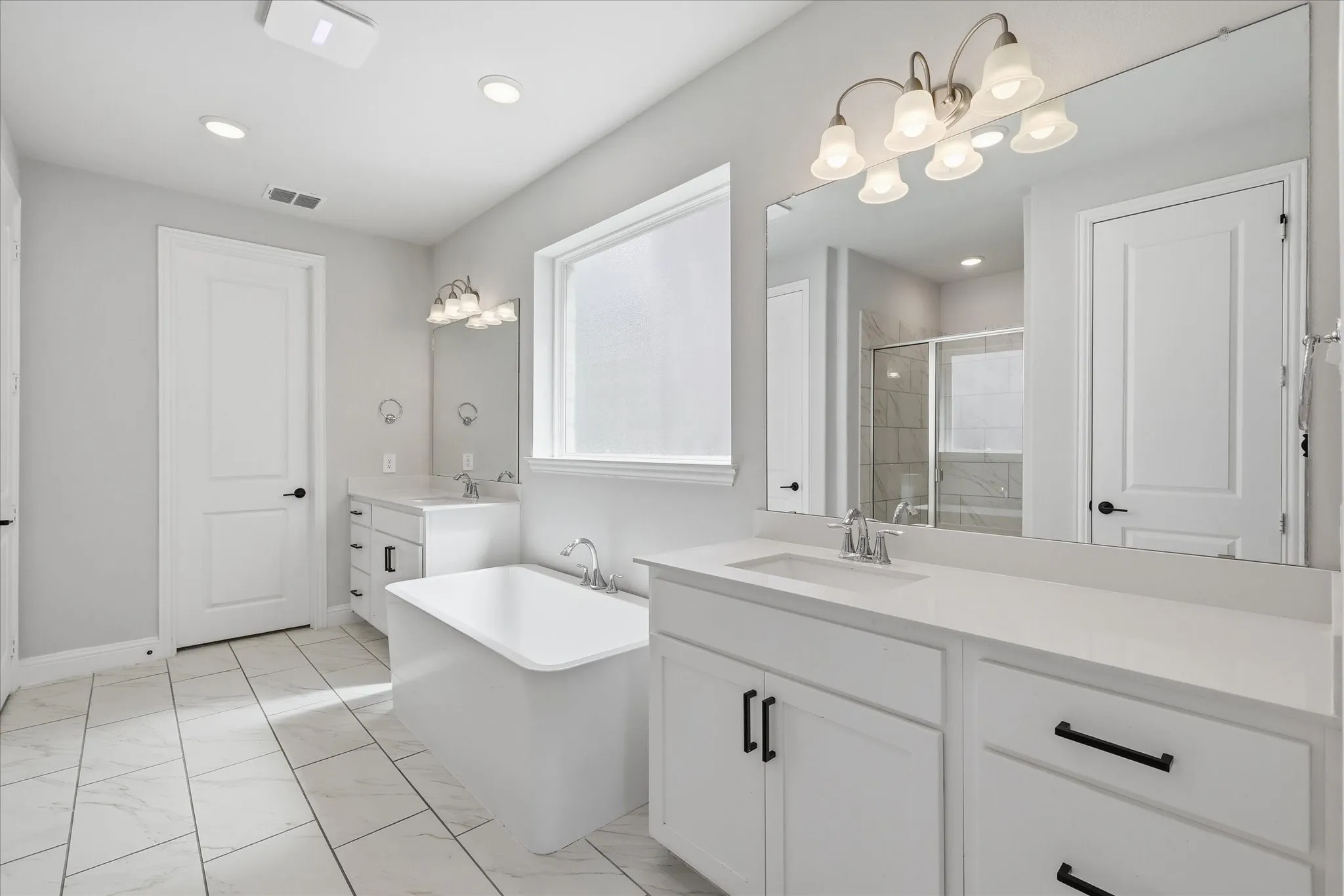 Spacious bathroom featuring a freestanding soaking tub, dual vanities with white cabinetry, and large format tile flooring
