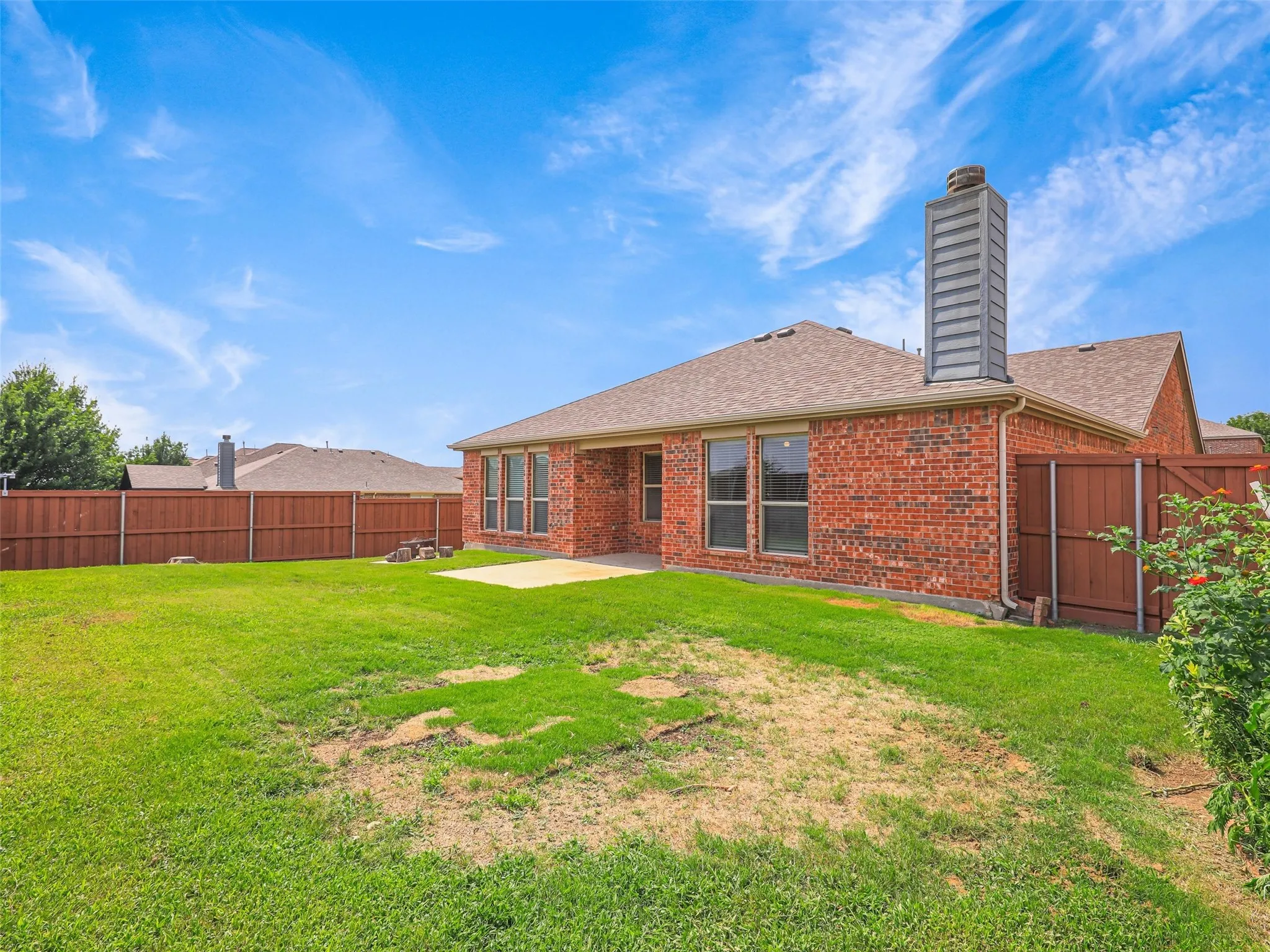 Spacious backyard with a privacy fence, concrete patio, brick exterior, multiple windows, and a prominent chimney