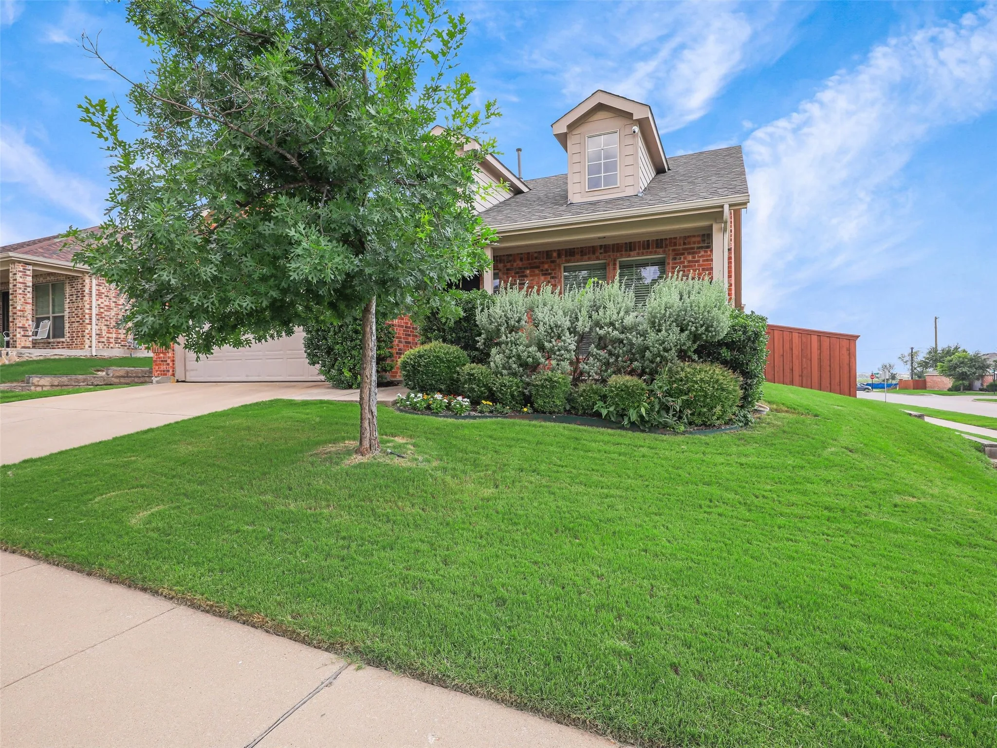 Brick facade residence featuring a front dormer, attached garage, concrete driveway, lush lawn, and mature landscaping