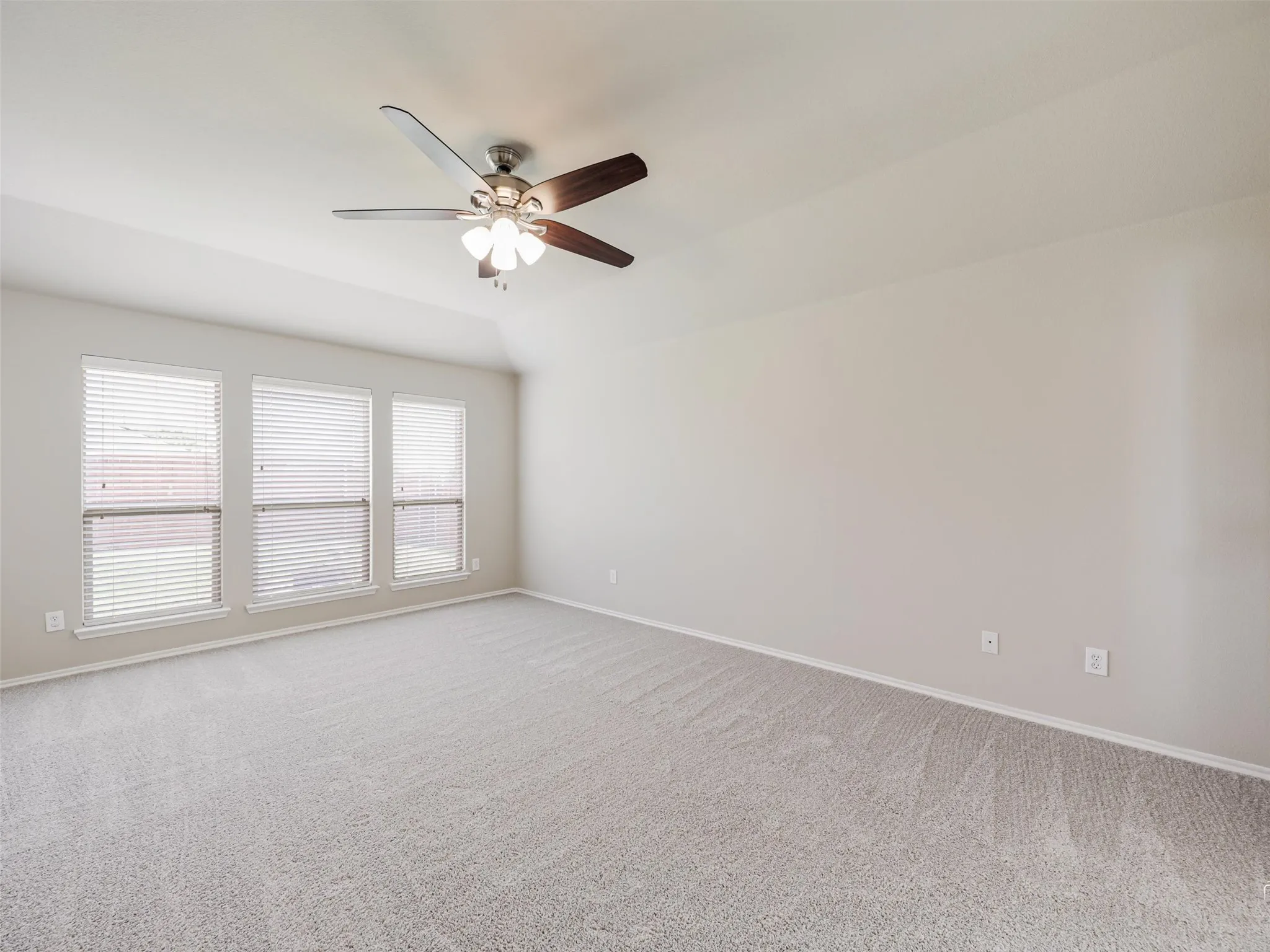 Spacious room featuring light-colored carpet, three windows with blinds, a vaulted ceiling, and a ceiling fan with integrated lighting