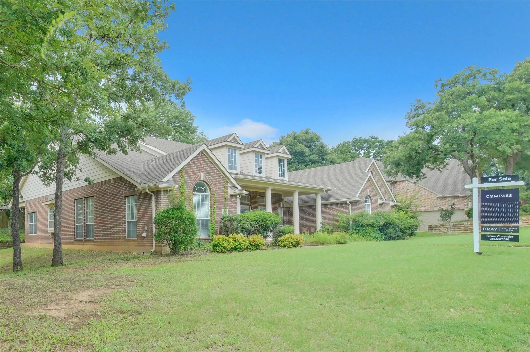 Brick exterior residence featuring a covered front porch, dormer windows, arched window accents, and mature landscaping