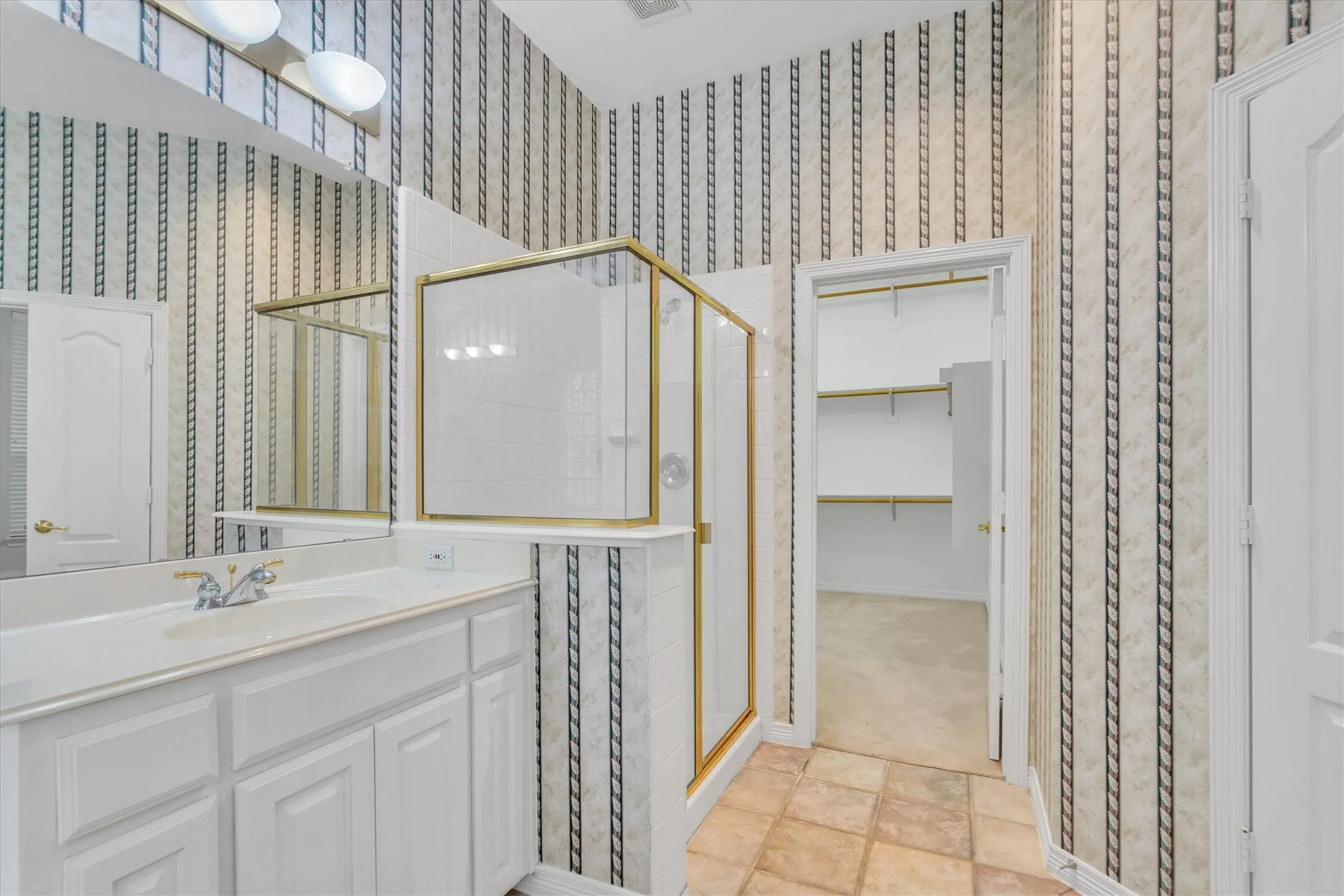 Primary Bathroom featuring a single vanity with integrated sink, gold-framed shower enclosure, patterned wallpaper, and a separate closet with shelving