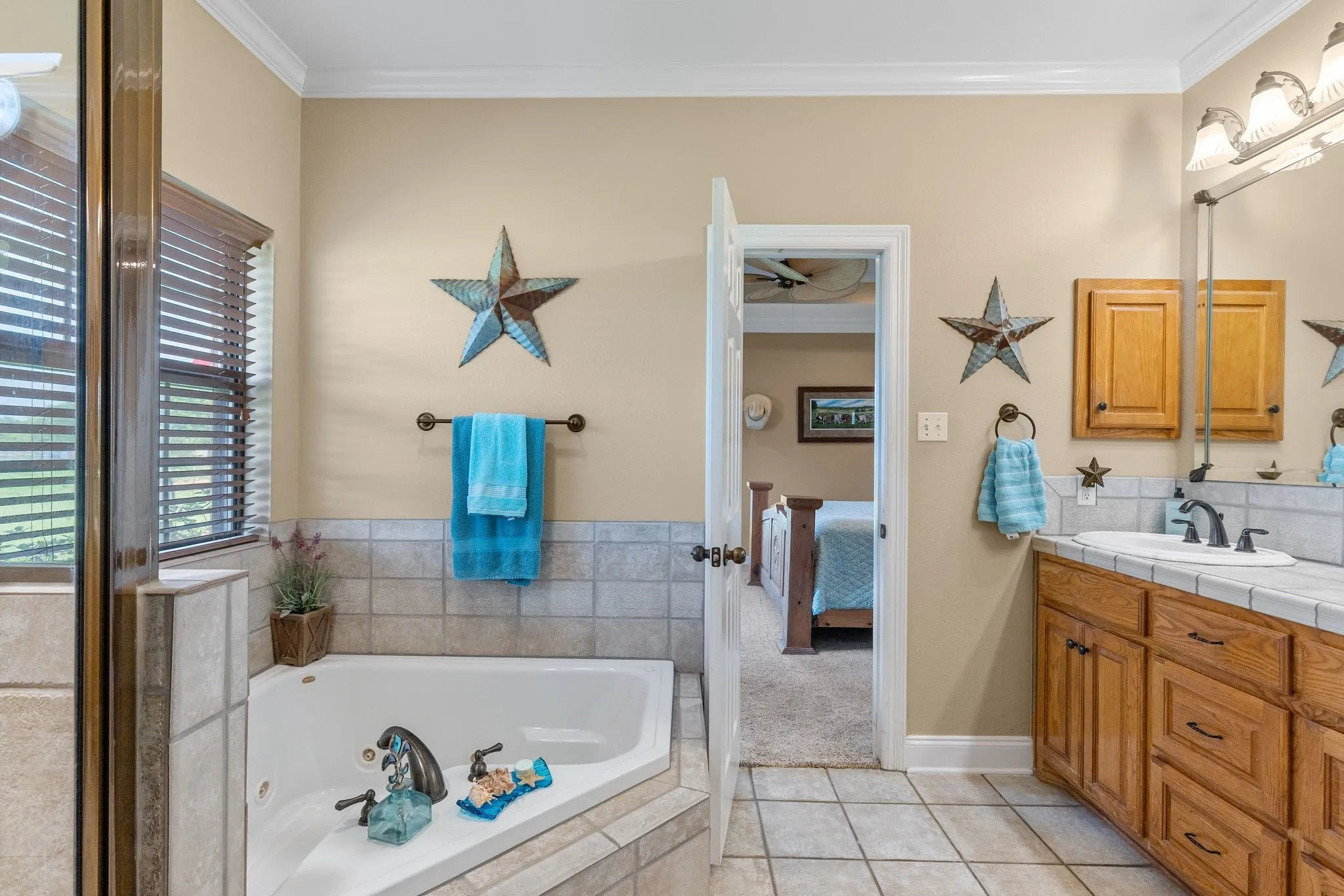 Bathroom featuring a corner soaking tub with surrounding tile, an adjacent glass-enclosed shower, a vanity with a tile countertop and wood-finish cabinetry, a window with blinds, and tile flooring