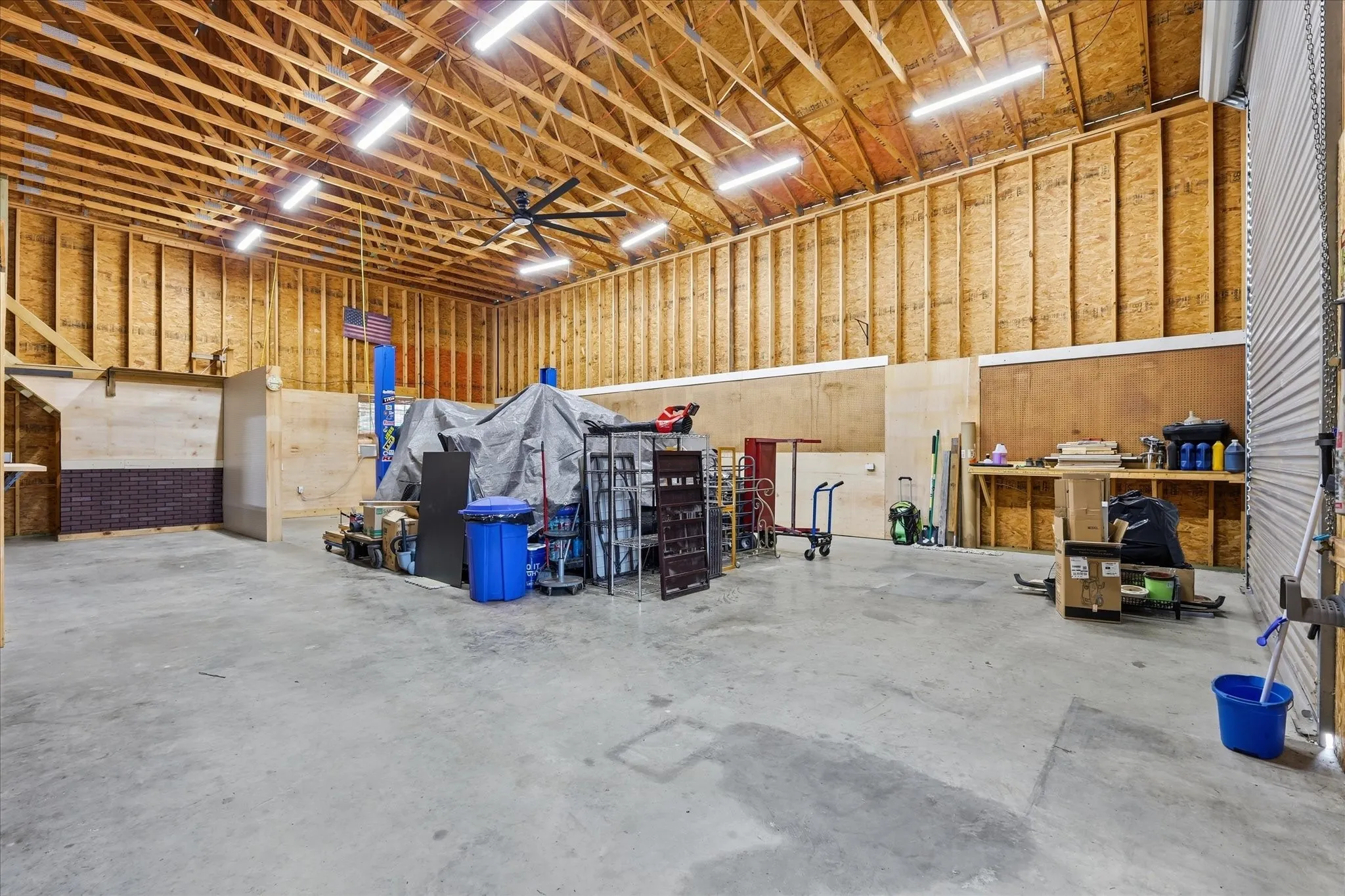 Spacious workshop area featuring exposed ceiling trusses, a concrete floor, built-in shelving, a large ceiling fan, and multiple overhead light fixtures