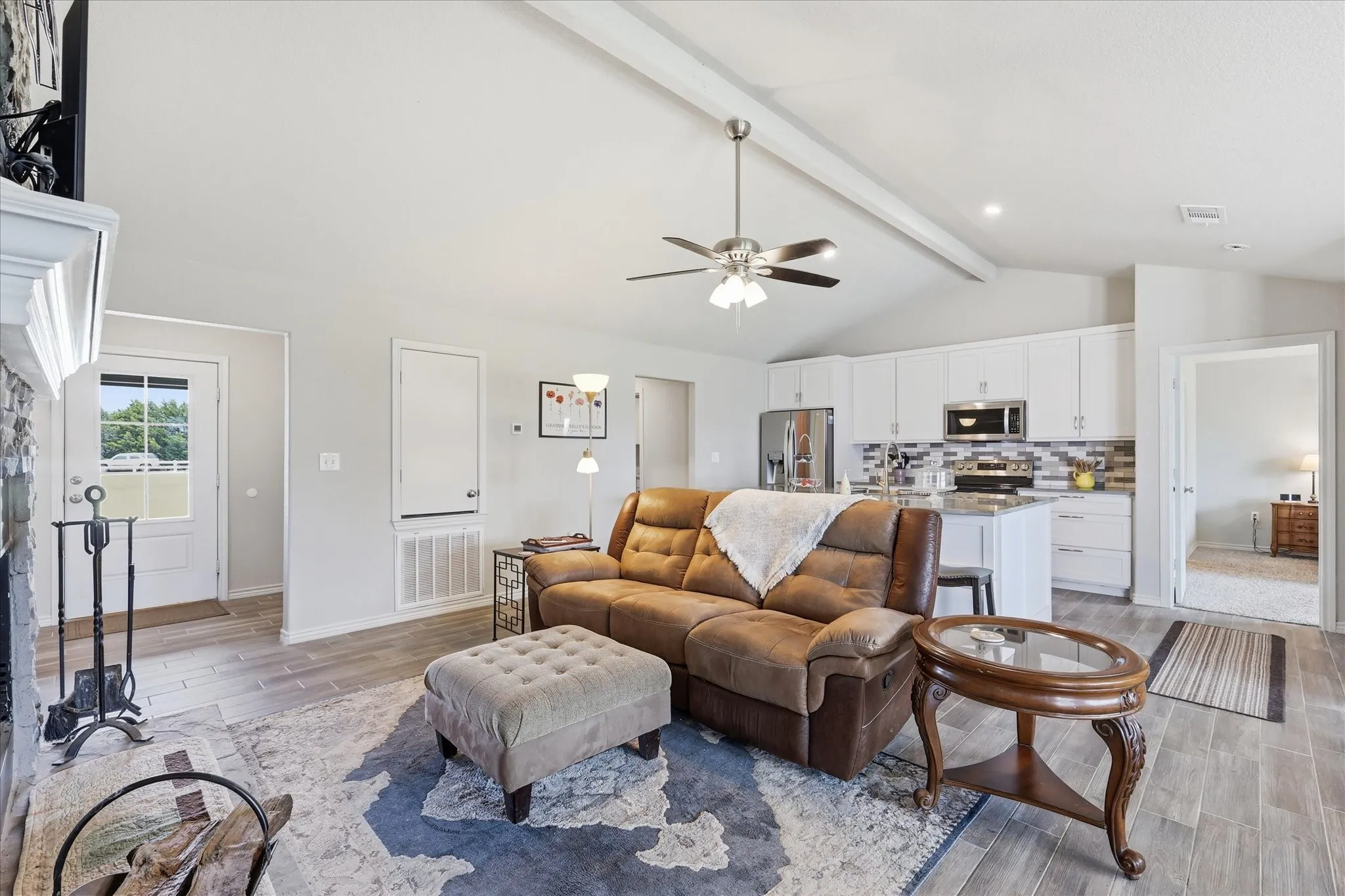Spacious living area featuring vaulted ceilings with exposed beams, wood-finish flooring, a stone-faced fireplace, and an open-concept kitchen with white cabinetry, stainless steel appliances, and a tiled backsplash