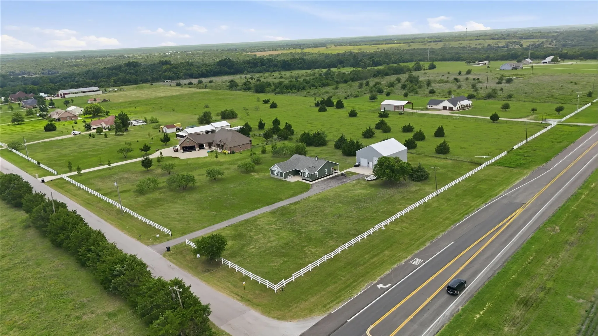 Expansive rural property featuring a single-story residence, a large detached workshop, a white picket fence, and a paved highway frontage