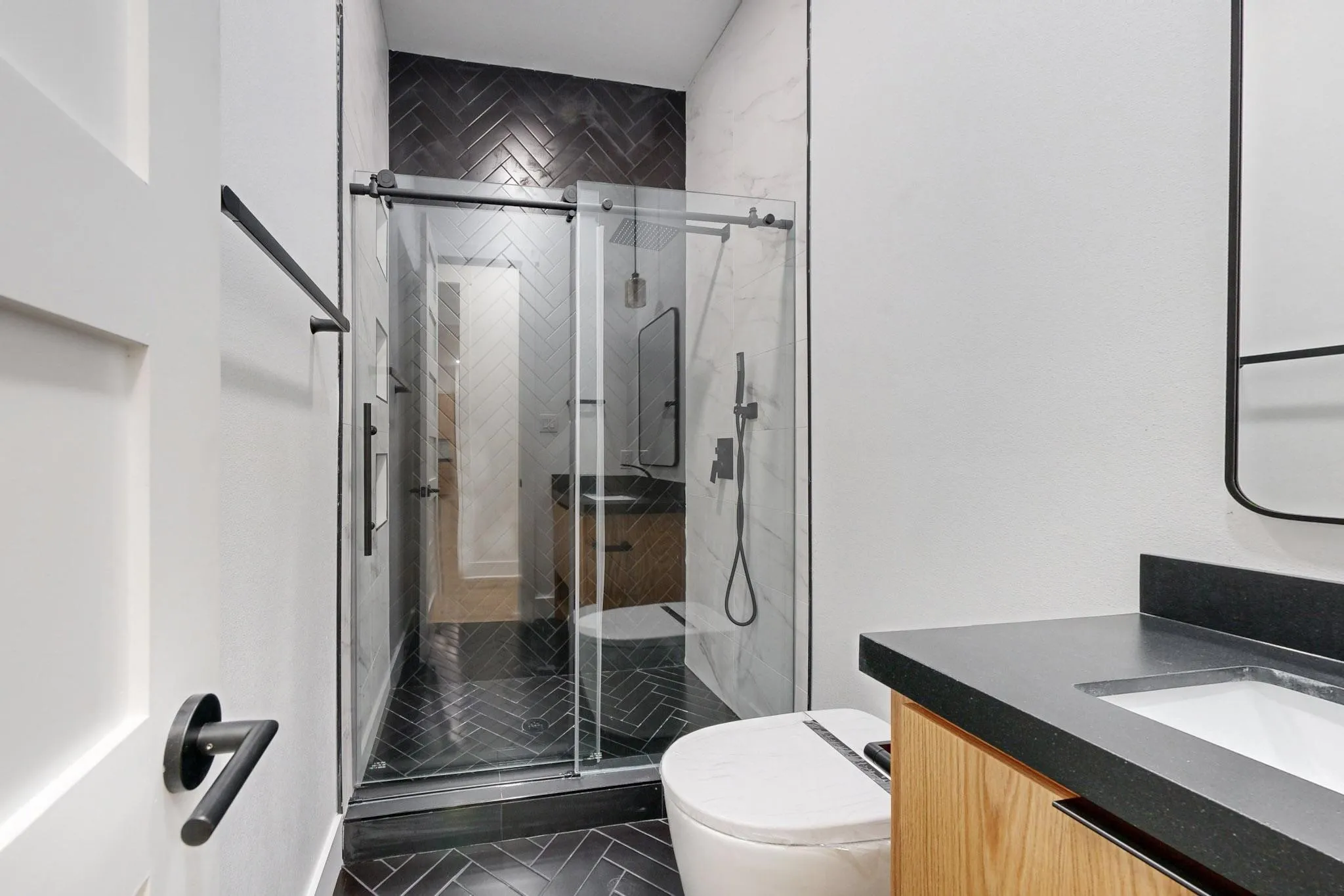 Contemporary bathroom featuring a glass-enclosed shower with black herringbone tile, a floating wood-finish vanity with a built-in sink and dark countertop, and matte black fixtures