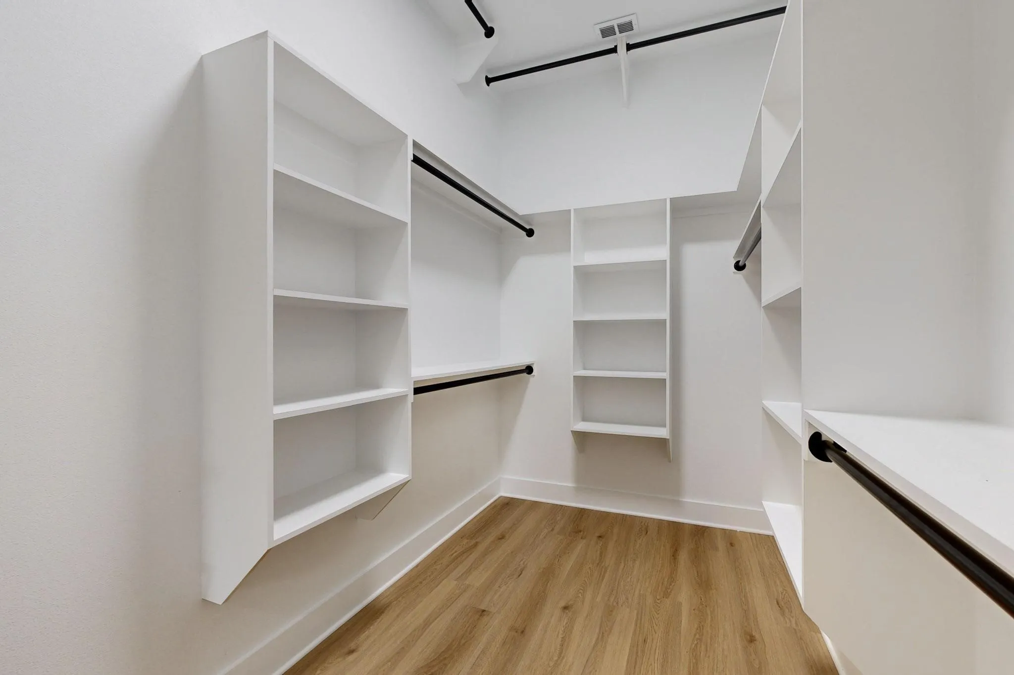 Walk-in closet featuring built-in white shelving, dark-finish hanging rods, and wood-finish flooring
