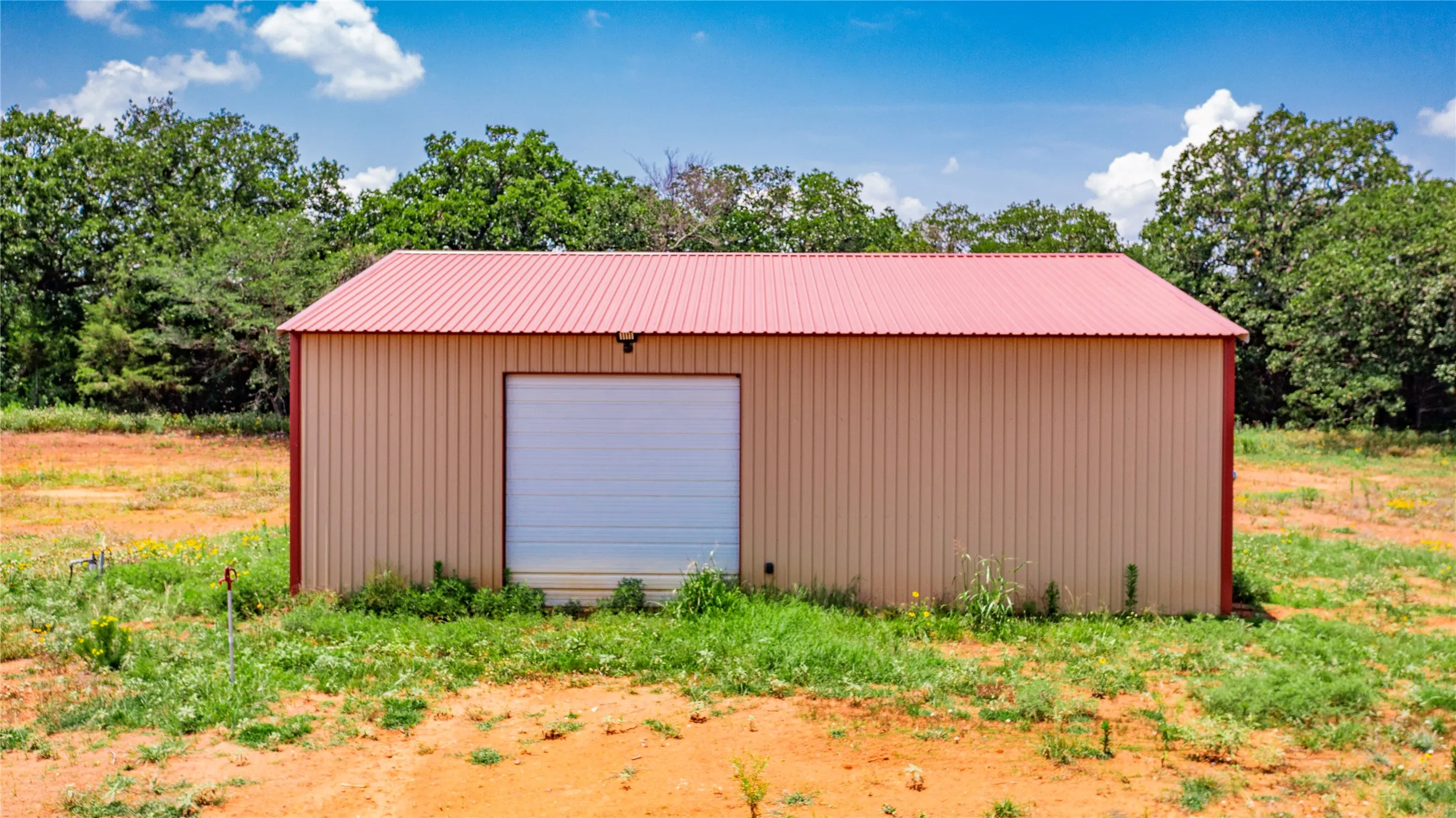Metal utility building featuring a corrugated red roof, tan siding, and a white roll-up door