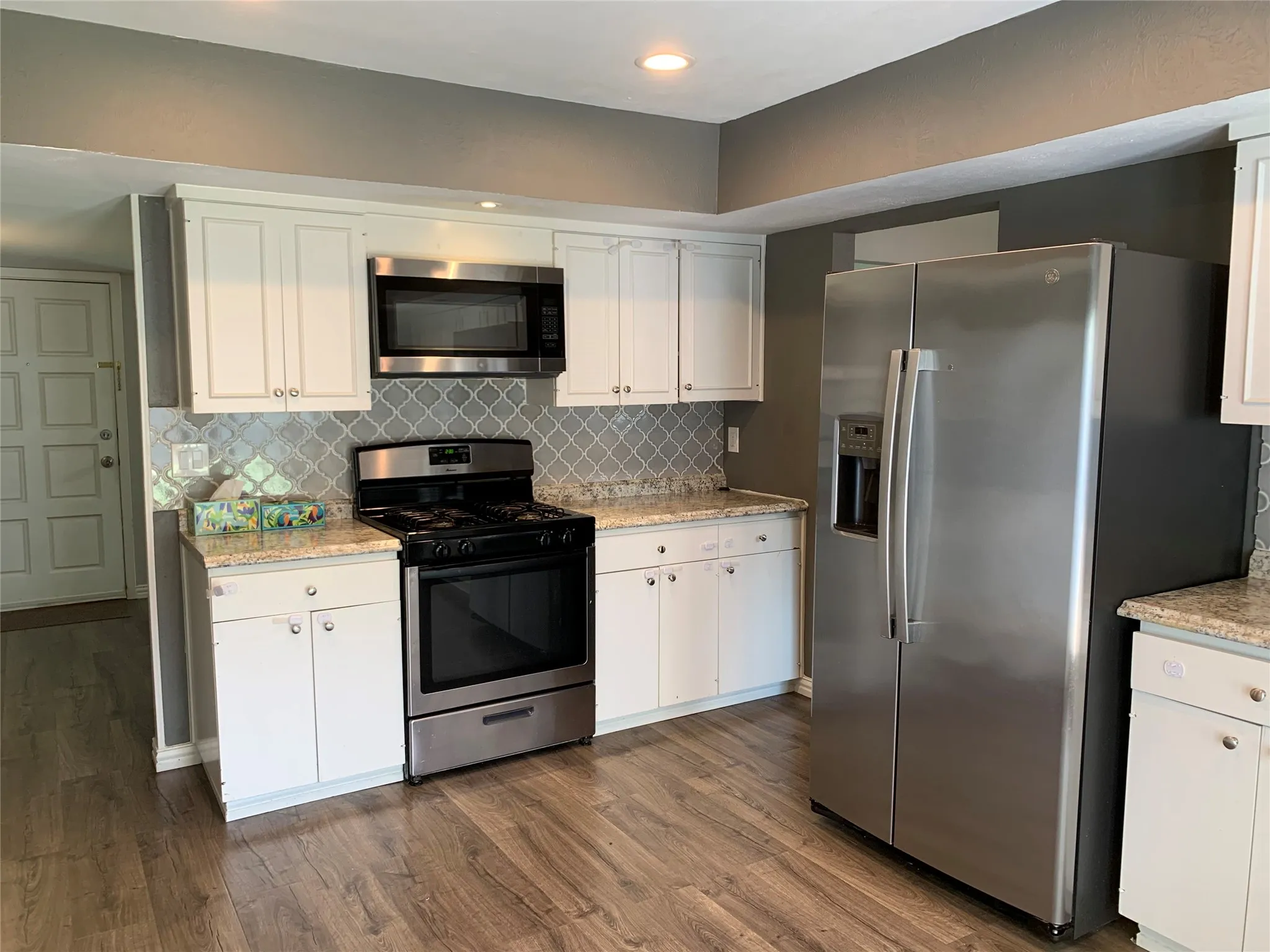 Modern kitchen featuring white cabinetry, light granite-finish countertops, stainless steel appliances, a stylish backsplash, and wood-finish flooring